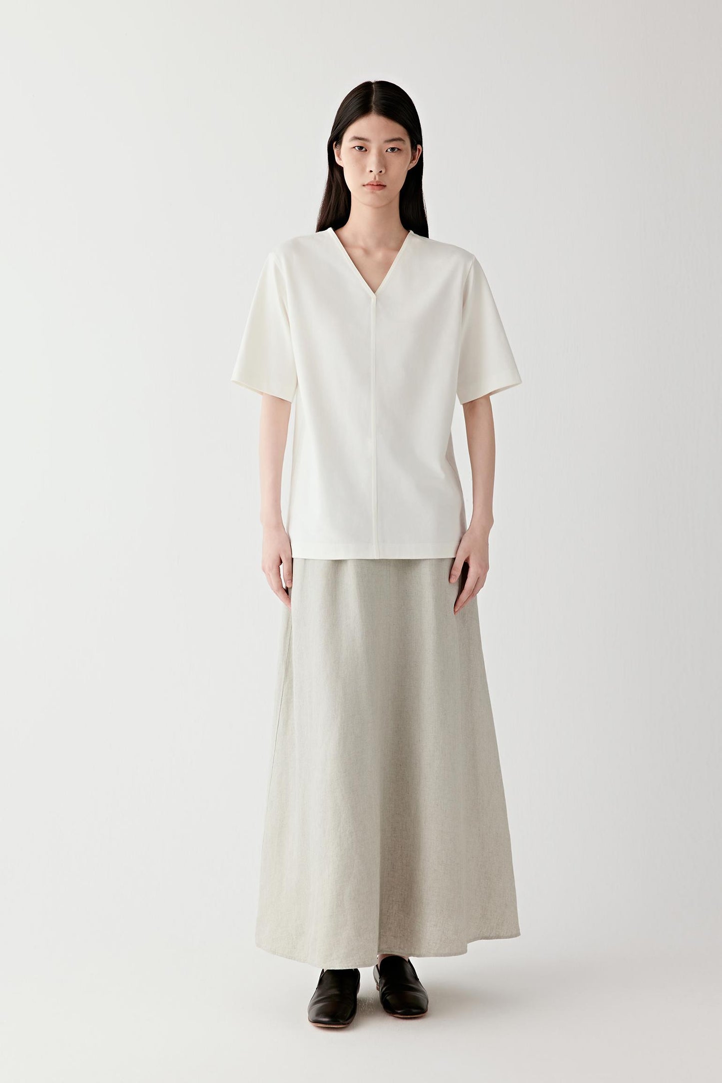 Full-length front view showing the relaxed silhouette, V-shaped neckline and short sleeves. The central seam construction introduces subtle structure while maintaining fluid movement through the garment.