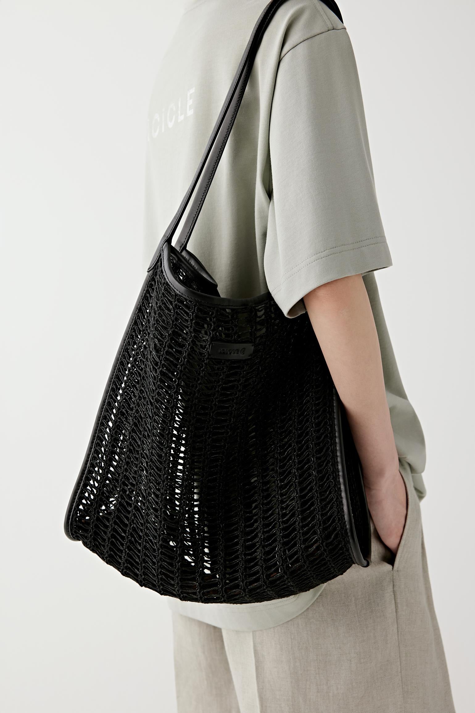 Side view of a woman carrying a black cotton mesh tote bag with leather straps, worn over a light outfit, emphasizing contrast between dark mesh and smooth leather