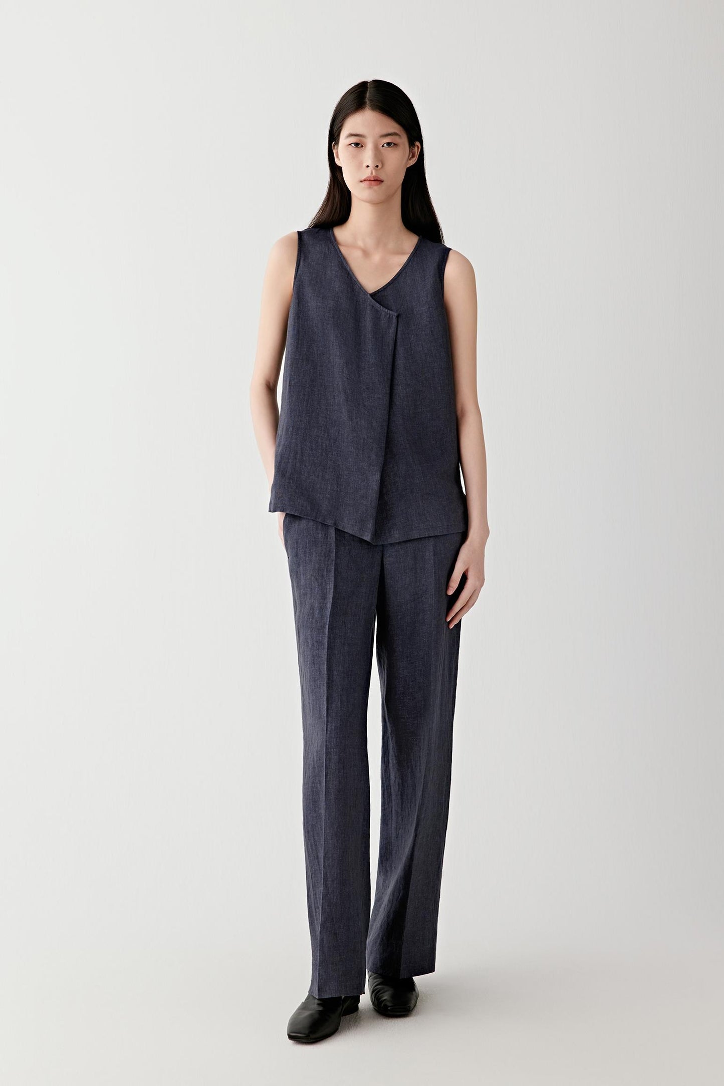 Full-length front view showing the long straight-leg silhouette shaped by front pleats and pressed leg folds. The dense linen weave maintains structure while allowing fluid movement.