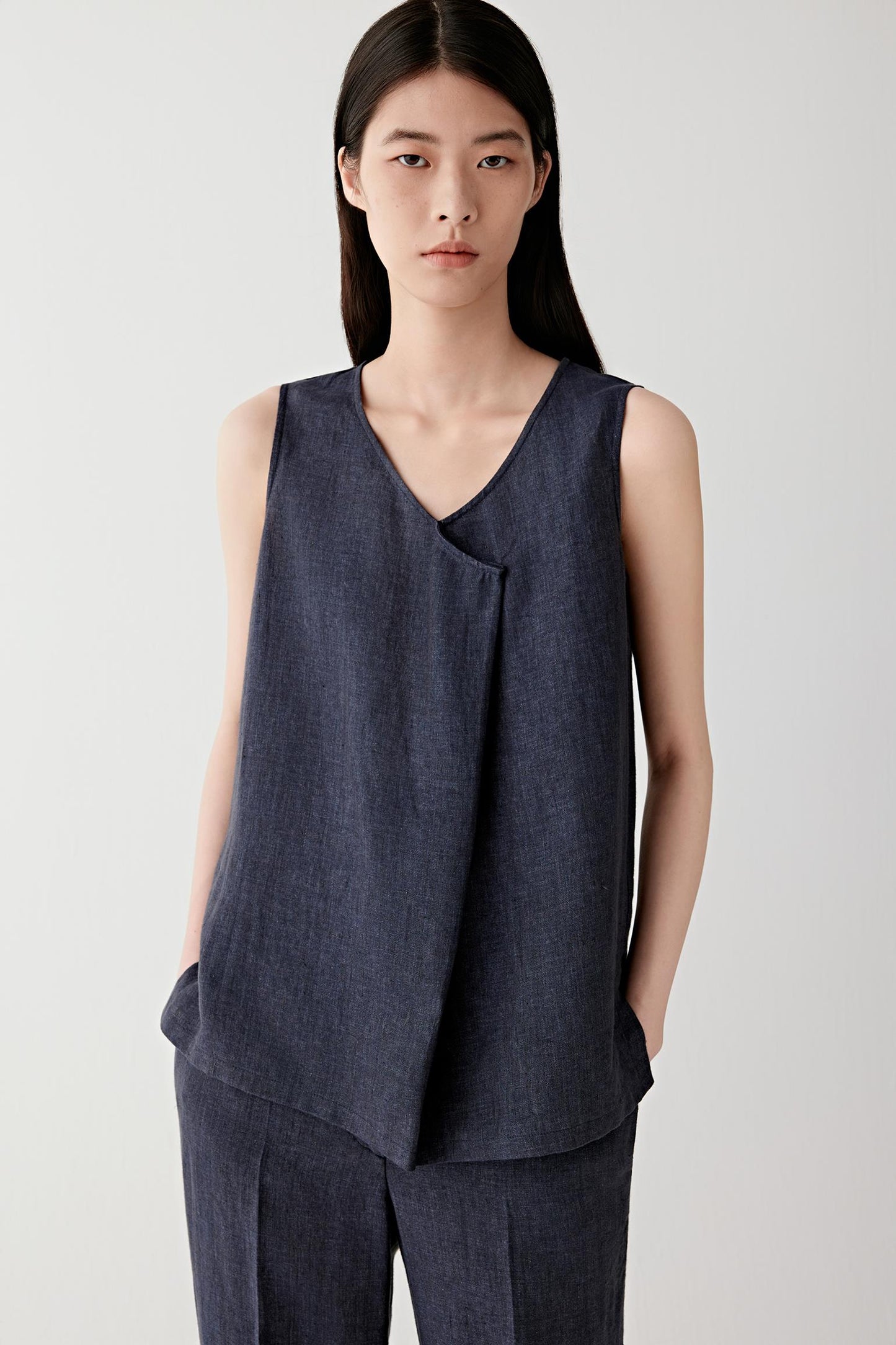 Front view of a woman wearing an indigo blue sleeveless blouse in linen, highlighting the lightweight texture, dry hand and subtle tonal variation across the matte surface. The woven linen structure creates a fluid yet softly structured silhouette.