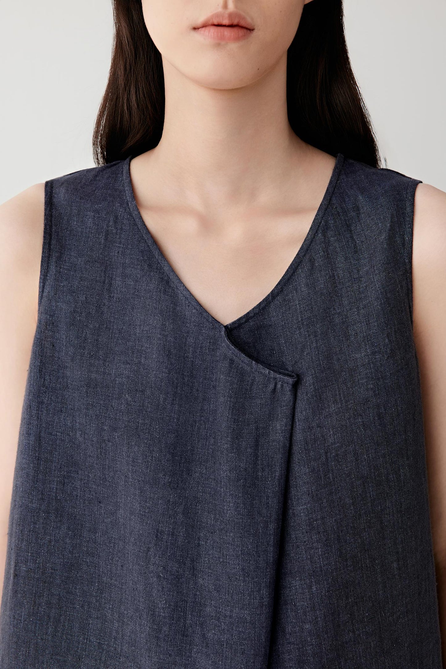Close-up portrait focusing on the V-shaped neckline and overlapping front panel detailing, revealing the dry woven texture and subtle tonal irregularities of the linen fabric.