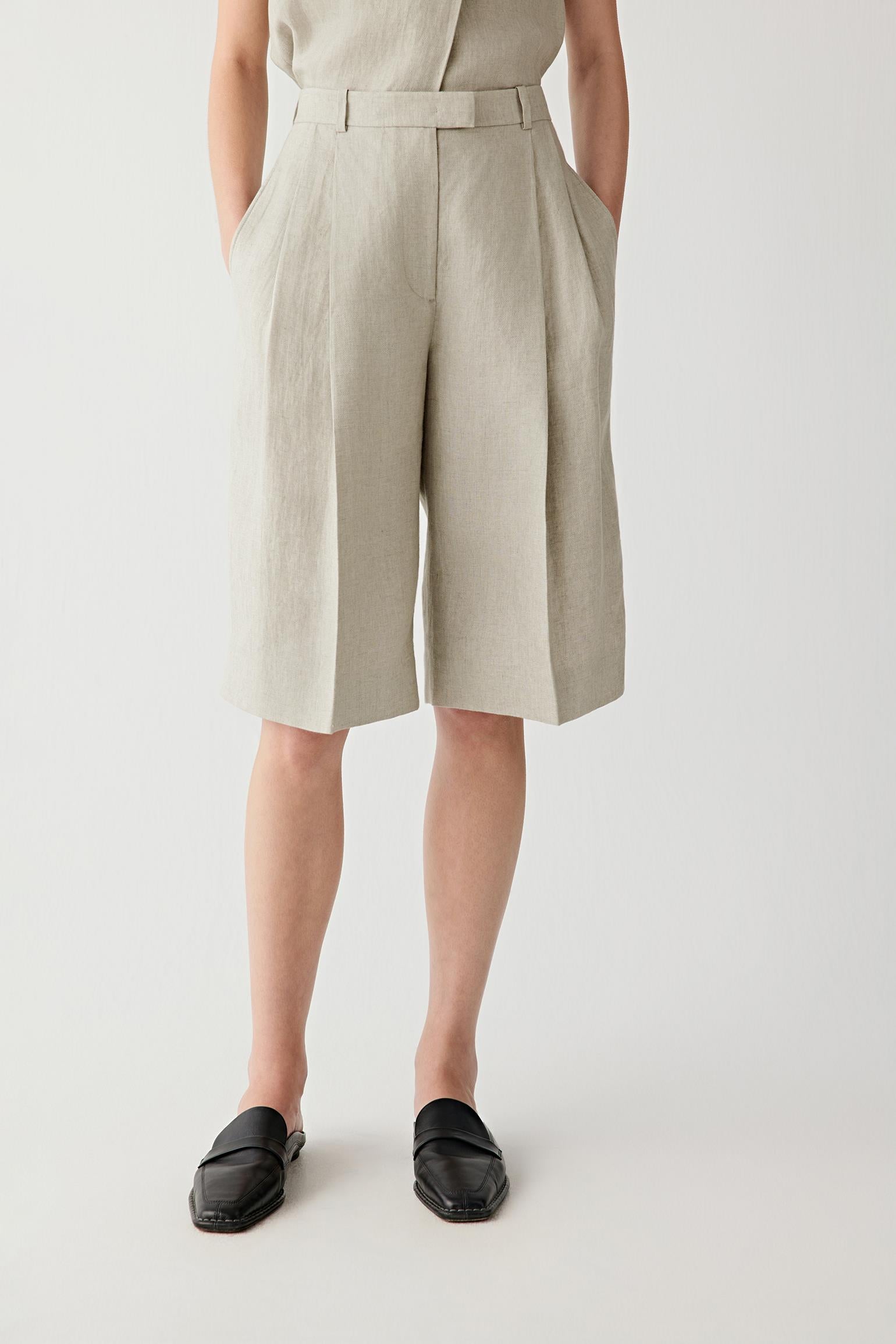Front view of a woman wearing beige-grey shorts in linen twill, highlighting the dry textured surface, visible grain and fluid drape of the woven fabric. The twill weave introduces subtle structure and tonal depth.