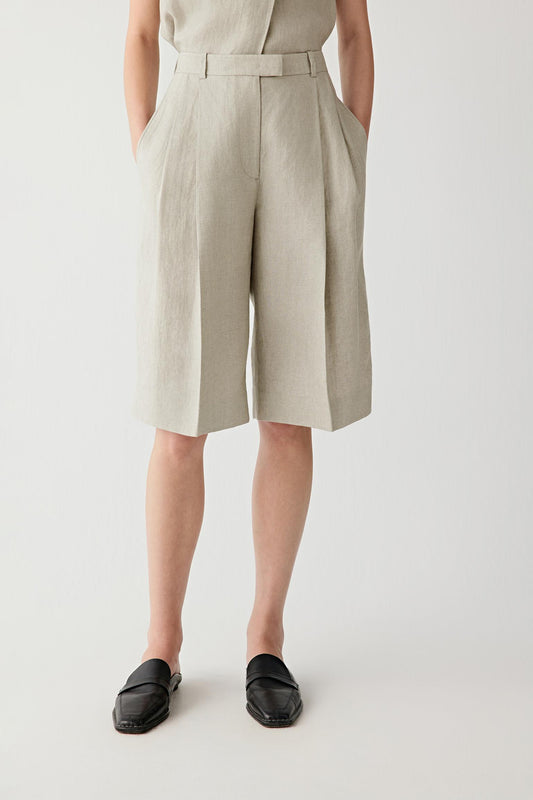 Front view of a woman wearing beige-grey shorts in linen twill, highlighting the dry textured surface, visible grain and fluid drape of the woven fabric. The twill weave introduces subtle structure and tonal depth.