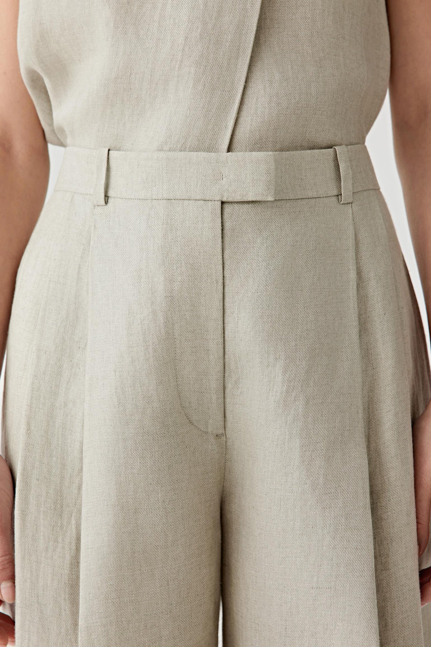 Close-up of the waistband and front pleat construction detailing the concealed fastening, belt loops and precise tailoring details. The woven twill texture remains visible across the surface.