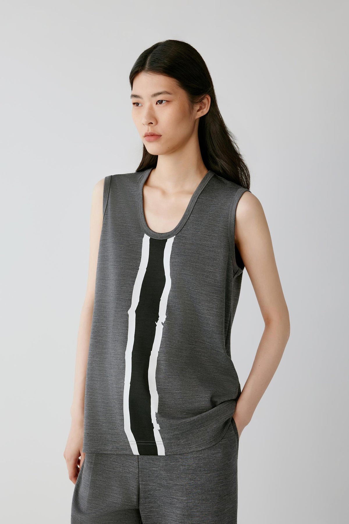 A woman modeling a sleeveless dark gray athletic top with a vertical black and white stripe down the front, paired with matching gray athletic pants, against a light gray studio background.