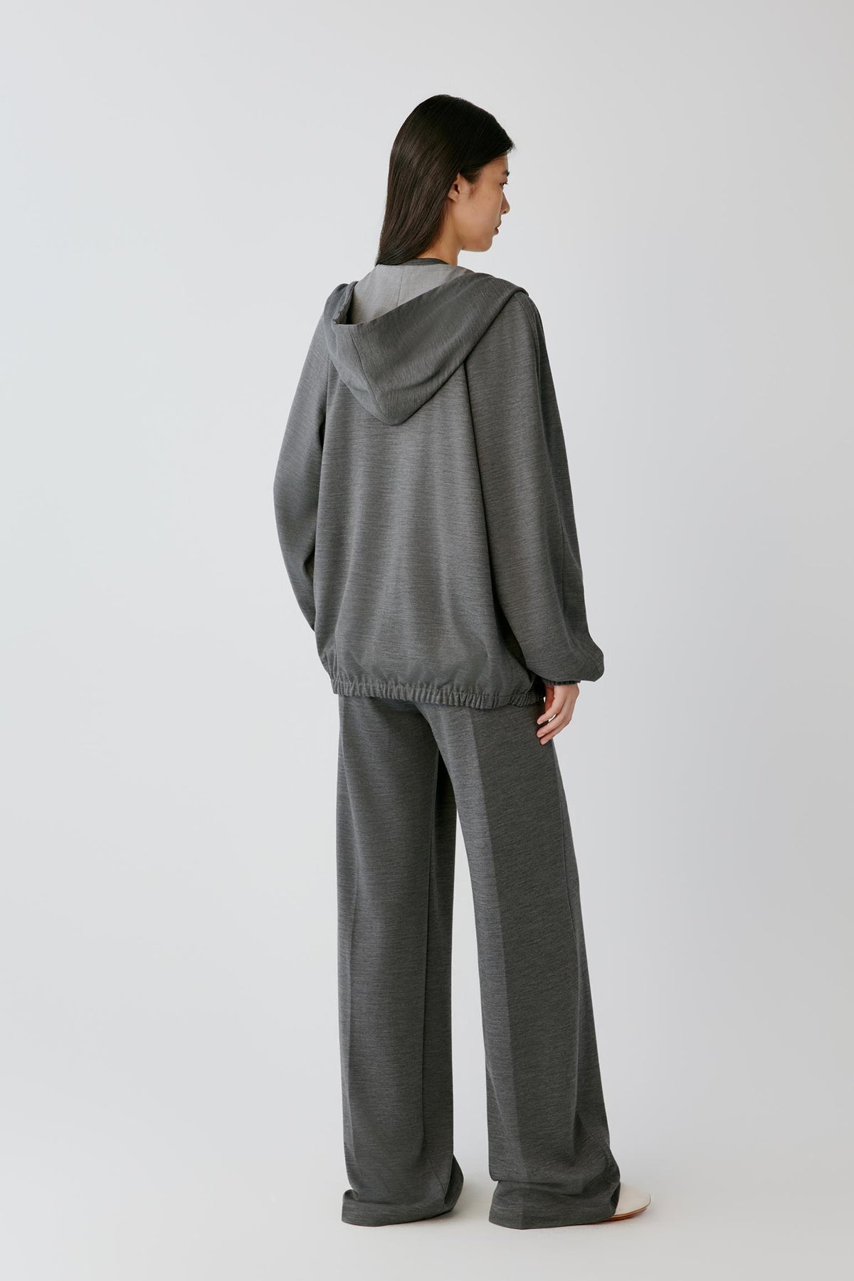 A woman shown from the back wearing a dark gray hooded sweatshirt and matching wide-leg pants against a light gray studio background.