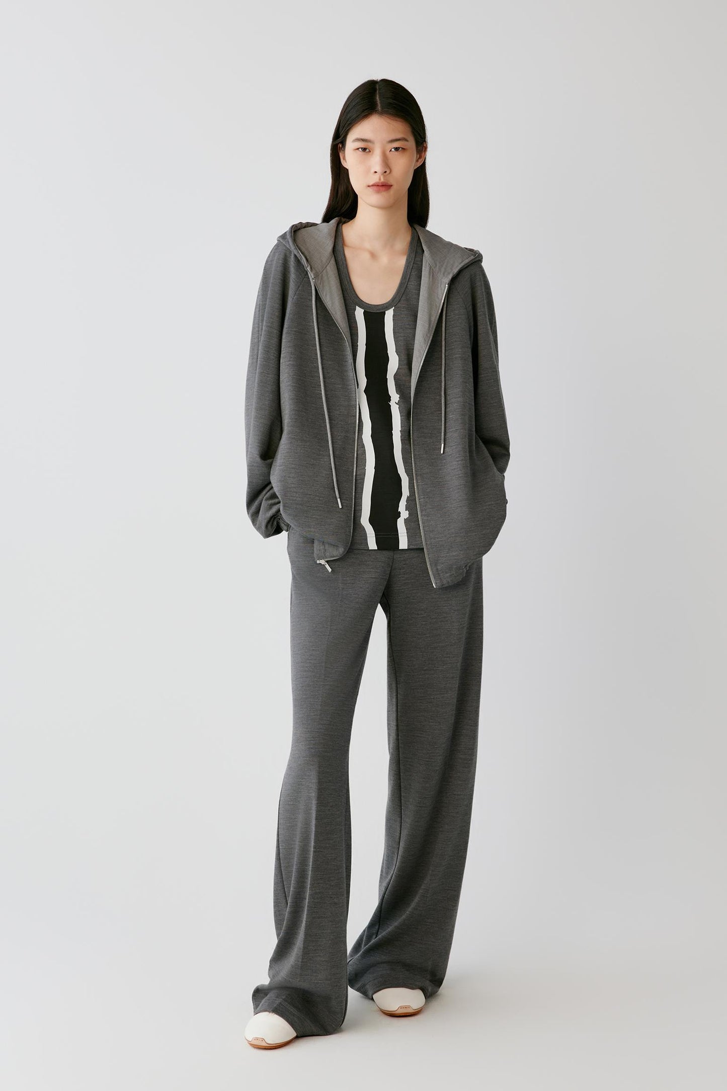 A model stands against a light gray studio background wearing a matching gray lounge set consisting of a zip-up hooded sweatshirt and wide-leg sweatpants, layered over a black and white striped tank top.