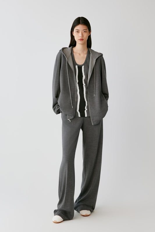 A model stands against a light gray studio background wearing a matching gray lounge set consisting of a zip-up hooded sweatshirt and wide-leg sweatpants, layered over a black and white striped tank top.