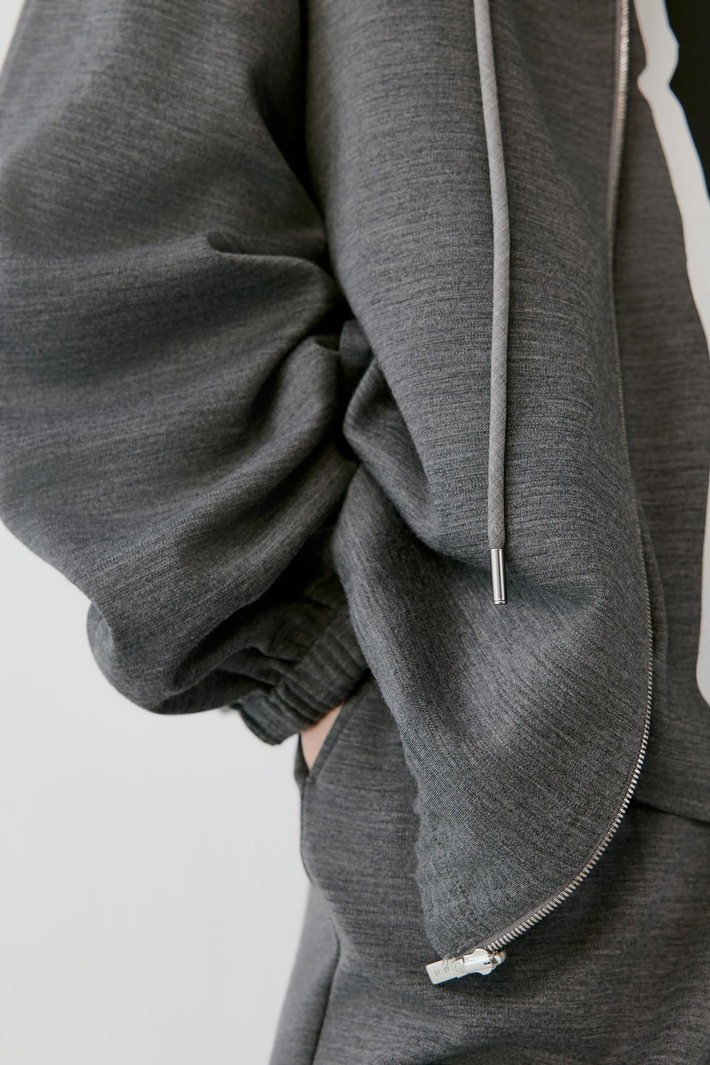 Close-up of a person wearing a dark gray heather zip-up hoodie with a drawstring and a white zipper accent, paired with matching gray sweatpants against a light gray background.