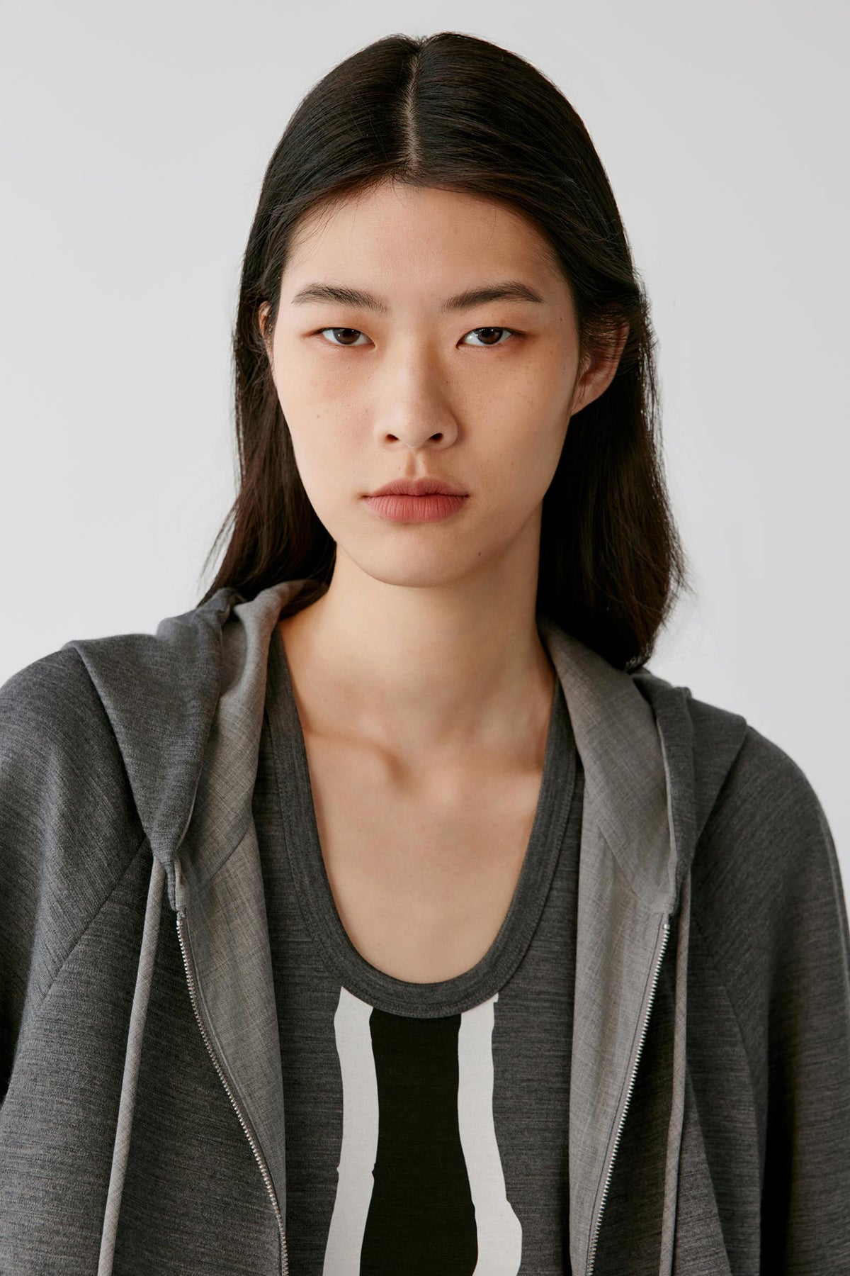 A woman with dark hair wearing a gray zip-up hoodie over a black and white striped shirt, photographed against a light gray background.