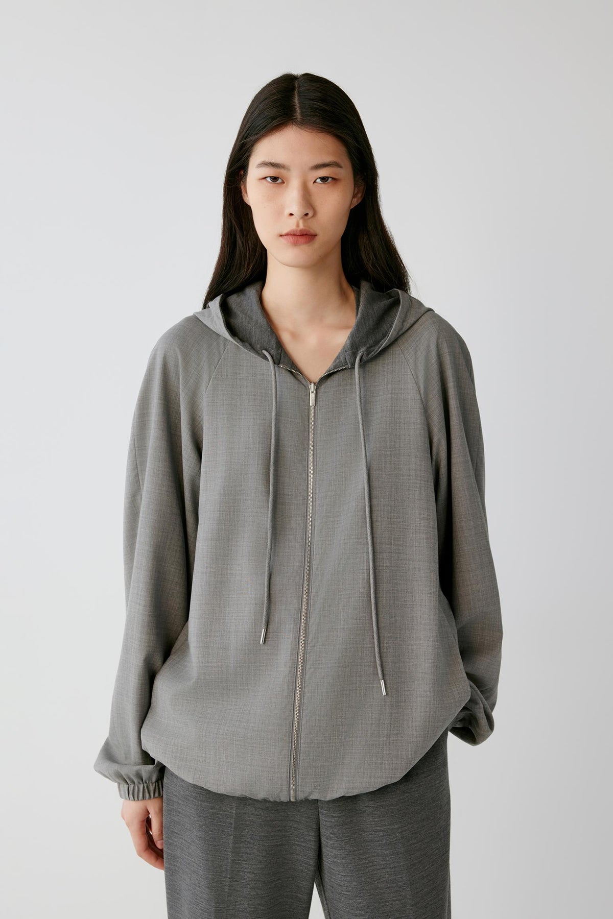 A model wearing a light gray zip-up hooded jacket with drawstrings and matching gray pants, standing against a plain light gray studio background.