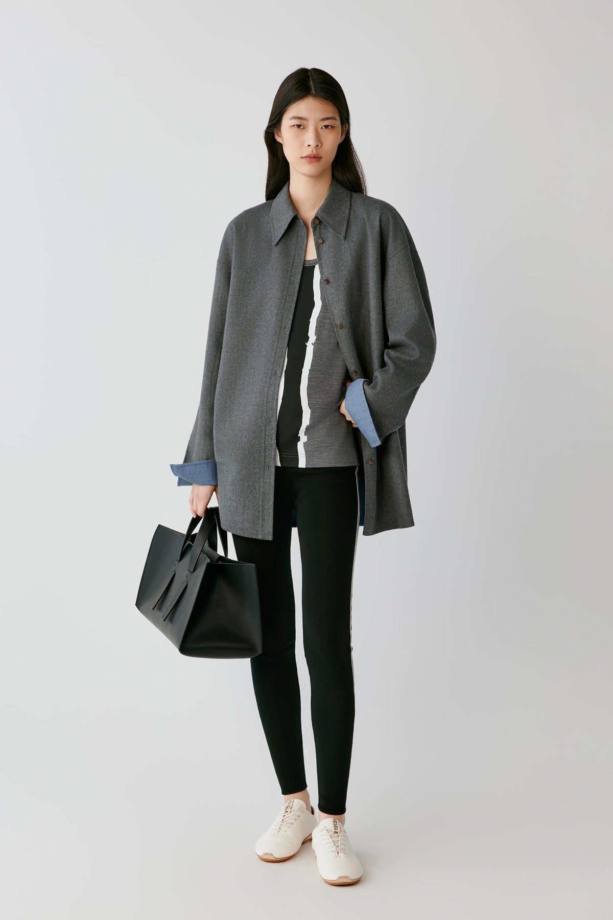 A model stands against a light gray studio background wearing an oversized dark gray button-up shirt over a black and white striped top, black leggings, and white sneakers, while holding a black tote bag.