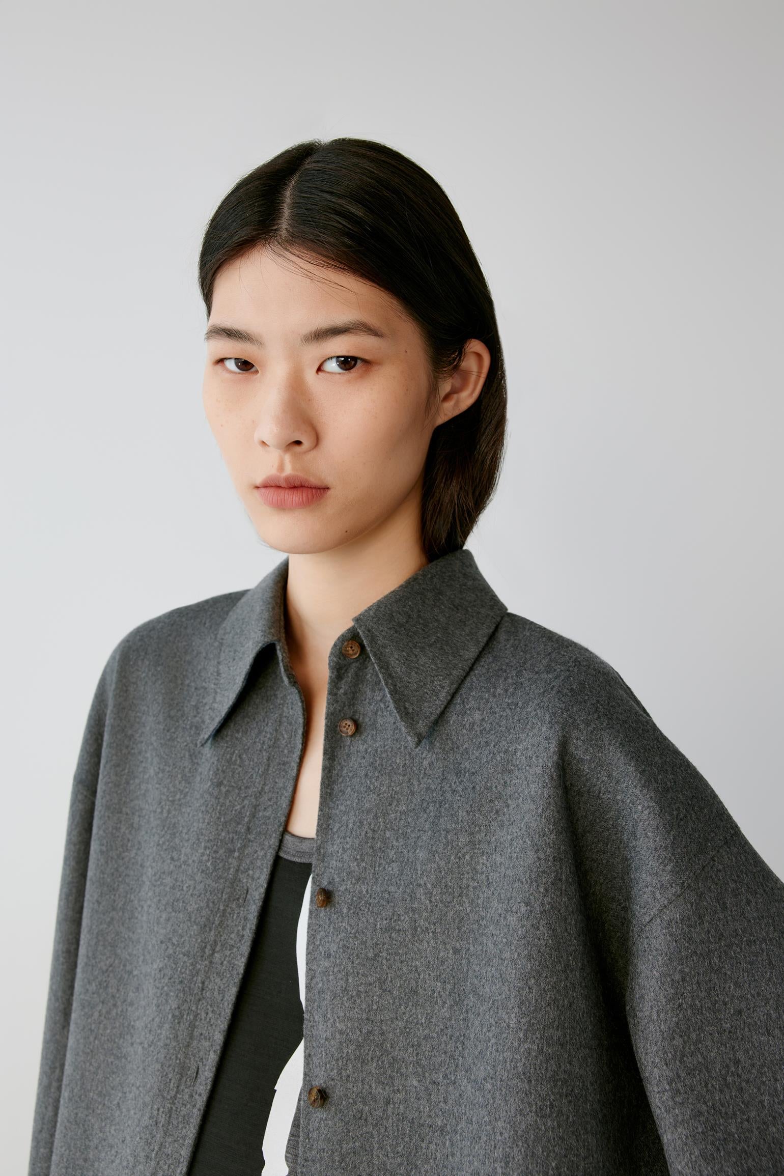 A model with dark hair wearing a gray button-down shirt jacket over a black and white top, photographed against a neutral light gray background.