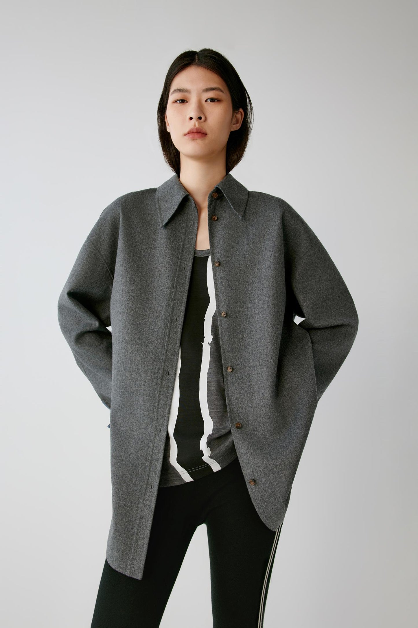 A model wearing a dark gray oversized button-up shirt jacket over a black and white striped top and black pants, standing against a light gray studio background.
