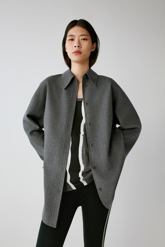 A model wearing a dark gray oversized button-up shirt jacket over a black and white striped top and black pants, standing against a light gray studio background.