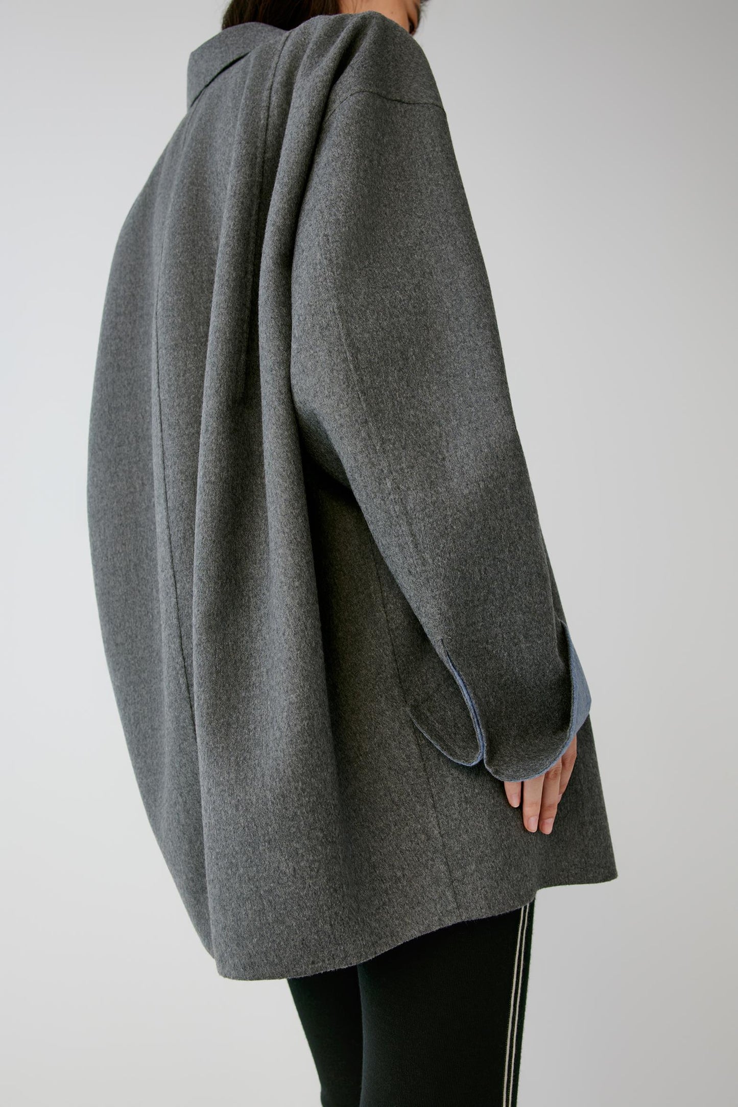 A person shown from the back wearing an oversized dark gray wool-blend coat with a relaxed, draped silhouette and black pants against a light gray studio background.