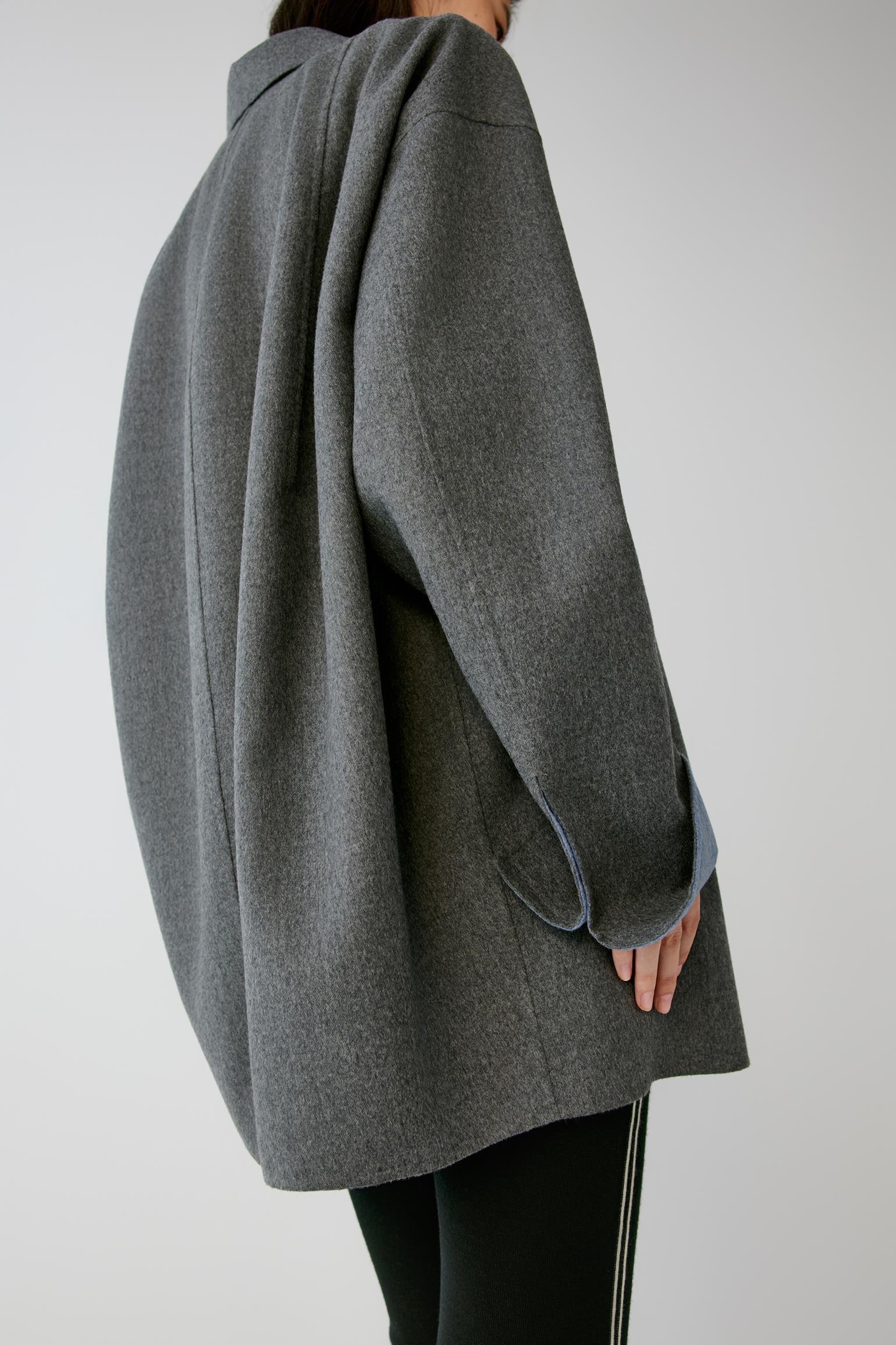 A person shown from the back wearing an oversized dark gray wool-blend coat with a relaxed, draped silhouette and black pants against a light gray studio background.