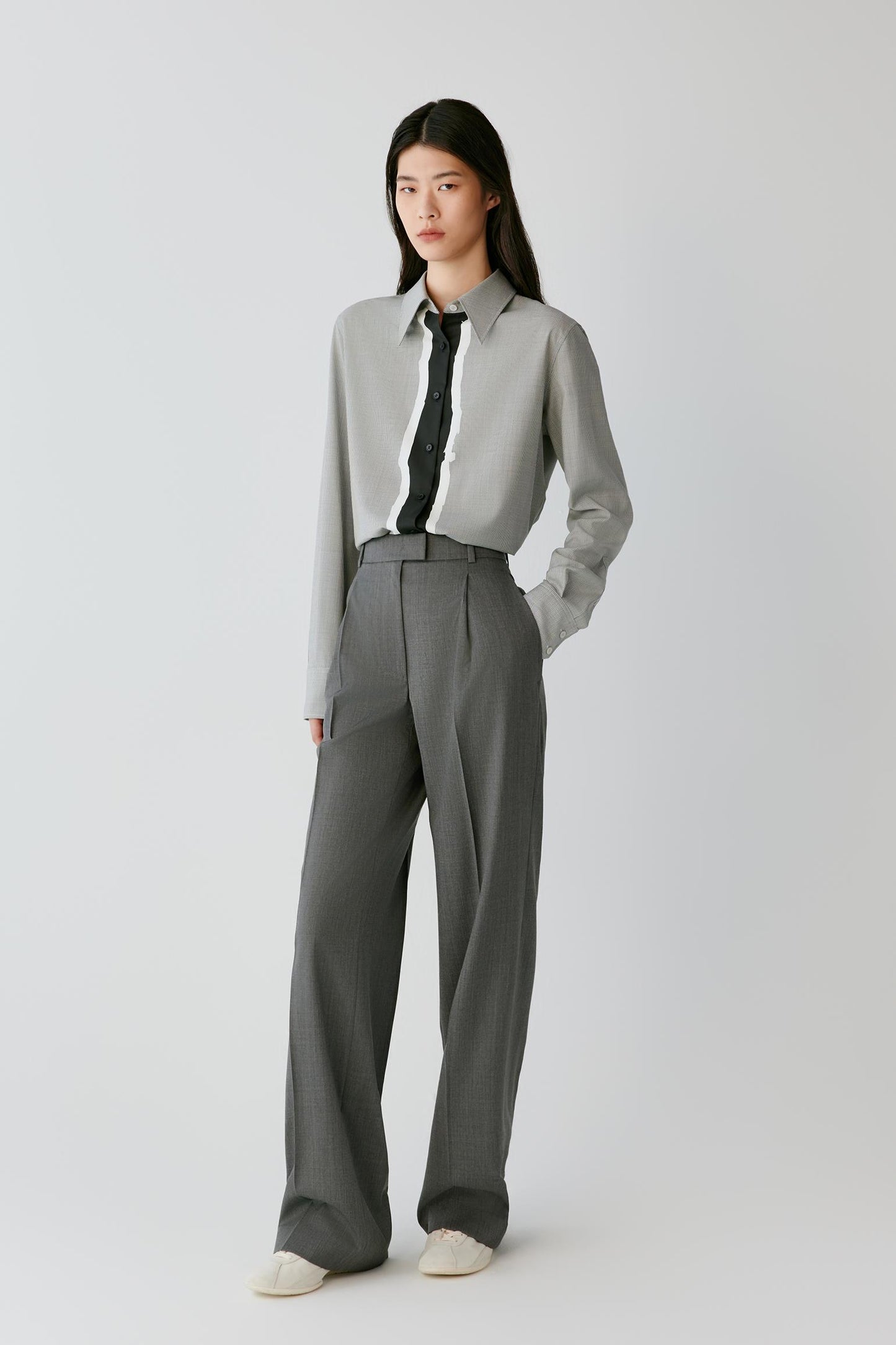 A model stands against a light gray studio background wearing a light gray long-sleeve button-up shirt, a black necktie, matching gray wide-leg trousers, and off-white slip-on shoes.