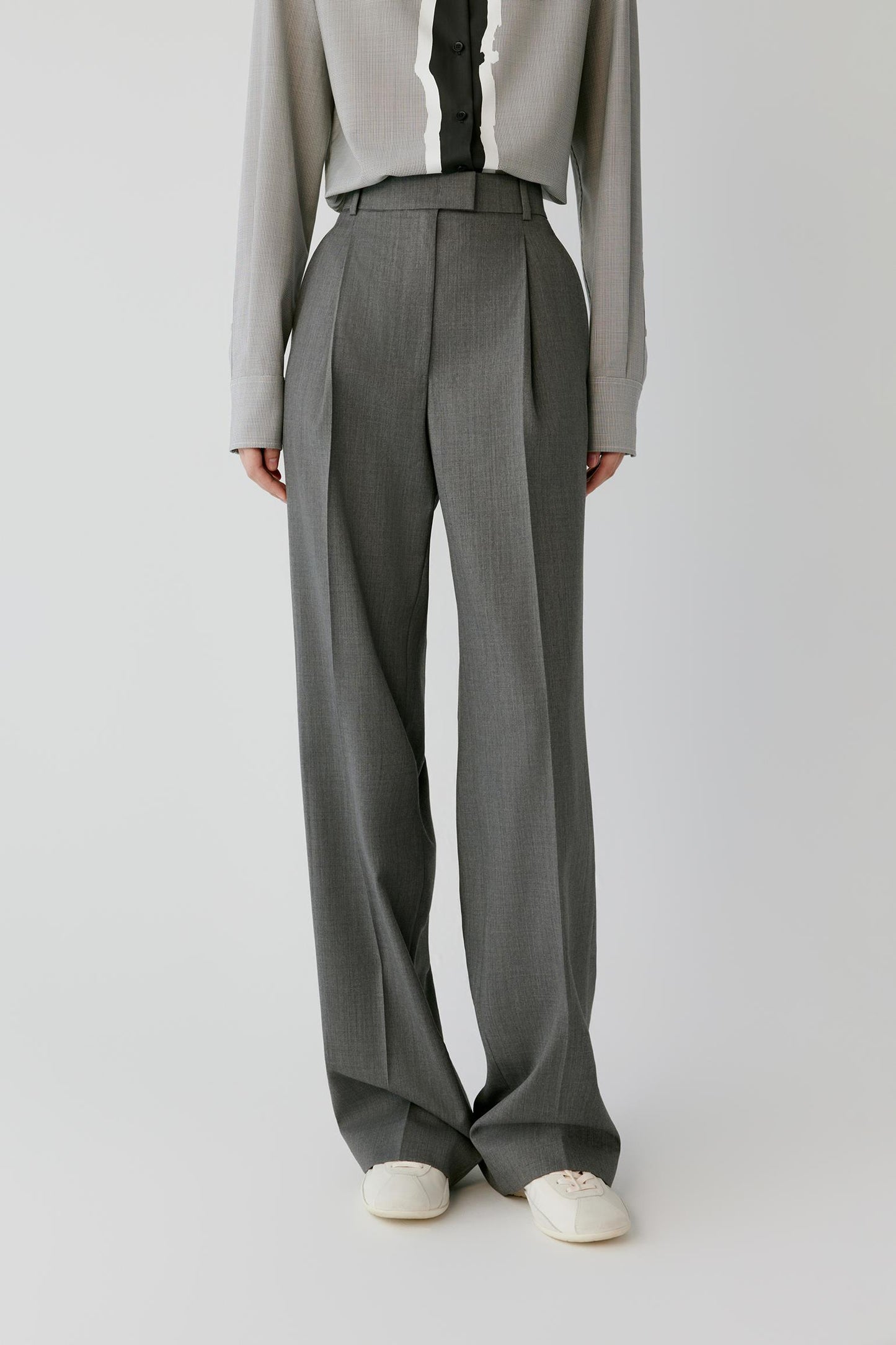 A model is shown from the chest down wearing high-waisted, wide-leg gray trousers with a matching belt, styled with a light gray shirt and white shoes against a plain light gray background.