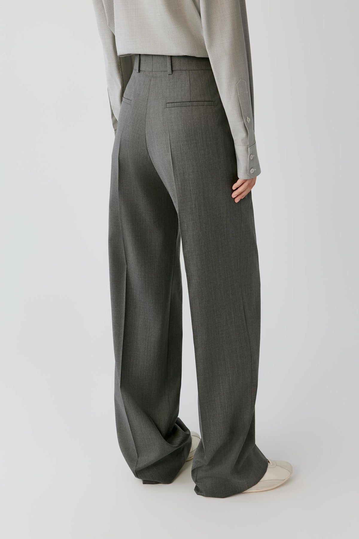 A person shown from the back wearing high-waisted, wide-leg gray trousers, a light gray long-sleeve top, and off-white slip-on shoes against a plain light gray studio background.