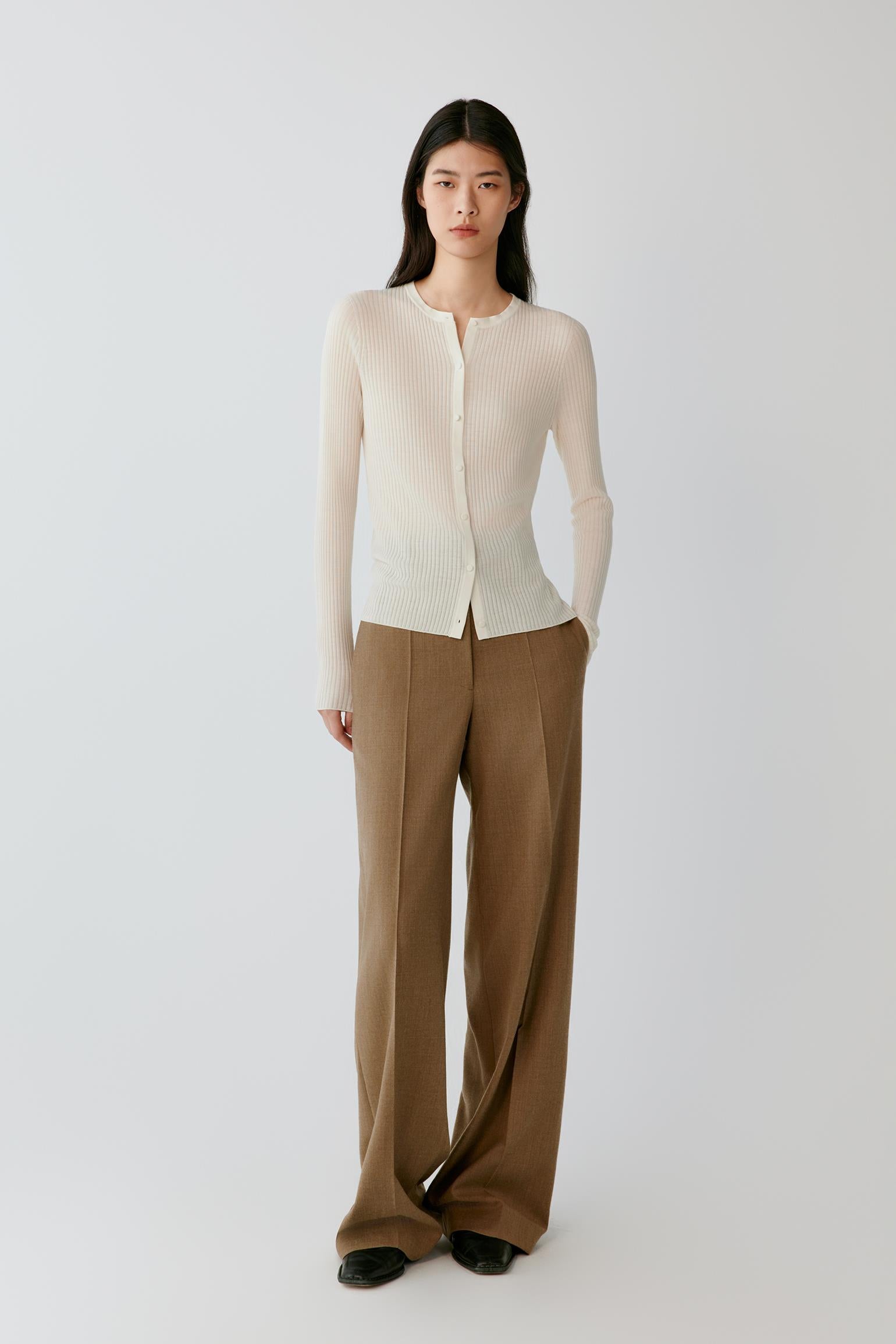 A model stands against a light gray studio background wearing a cream long-sleeve button-front knit top and wide-leg brown trousers.