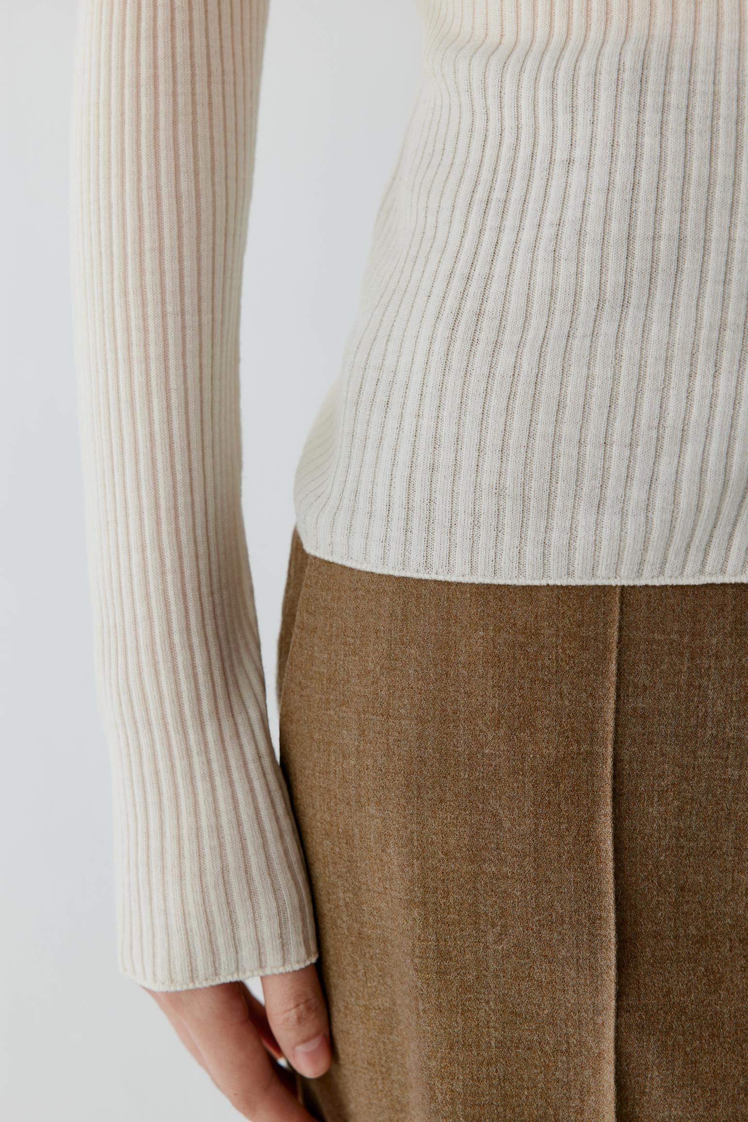 Close-up of a person wearing a ribbed cream long-sleeve top and brown textured skirt or trousers against a light neutral background.