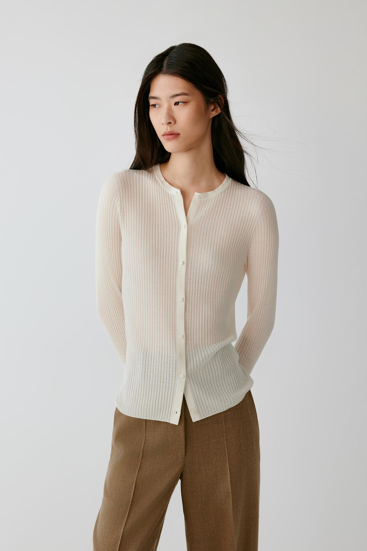 A woman modeling a cream ribbed knit cardigan with a buttoned front and long sleeves, paired with brown trousers, against a plain light gray studio background.