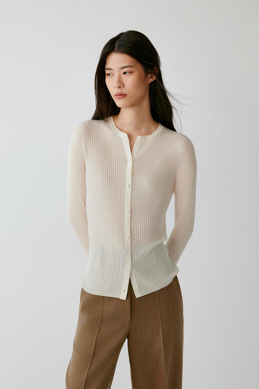 A woman modeling a cream ribbed knit cardigan with a buttoned front and long sleeves, paired with brown trousers, against a plain light gray studio background.