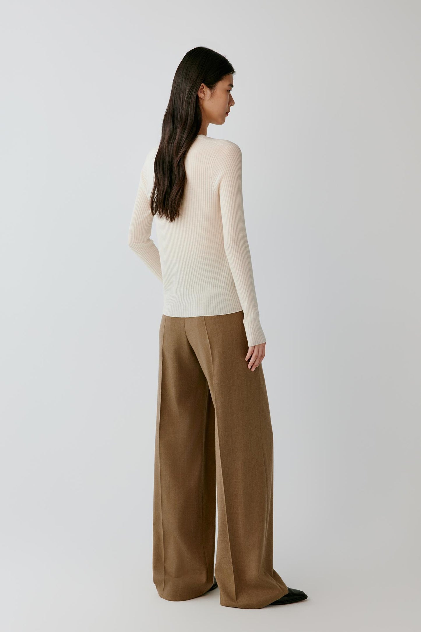 A woman is shown from the back wearing a cream-colored long-sleeve knit top and wide-leg brown trousers, standing against a light gray studio background.