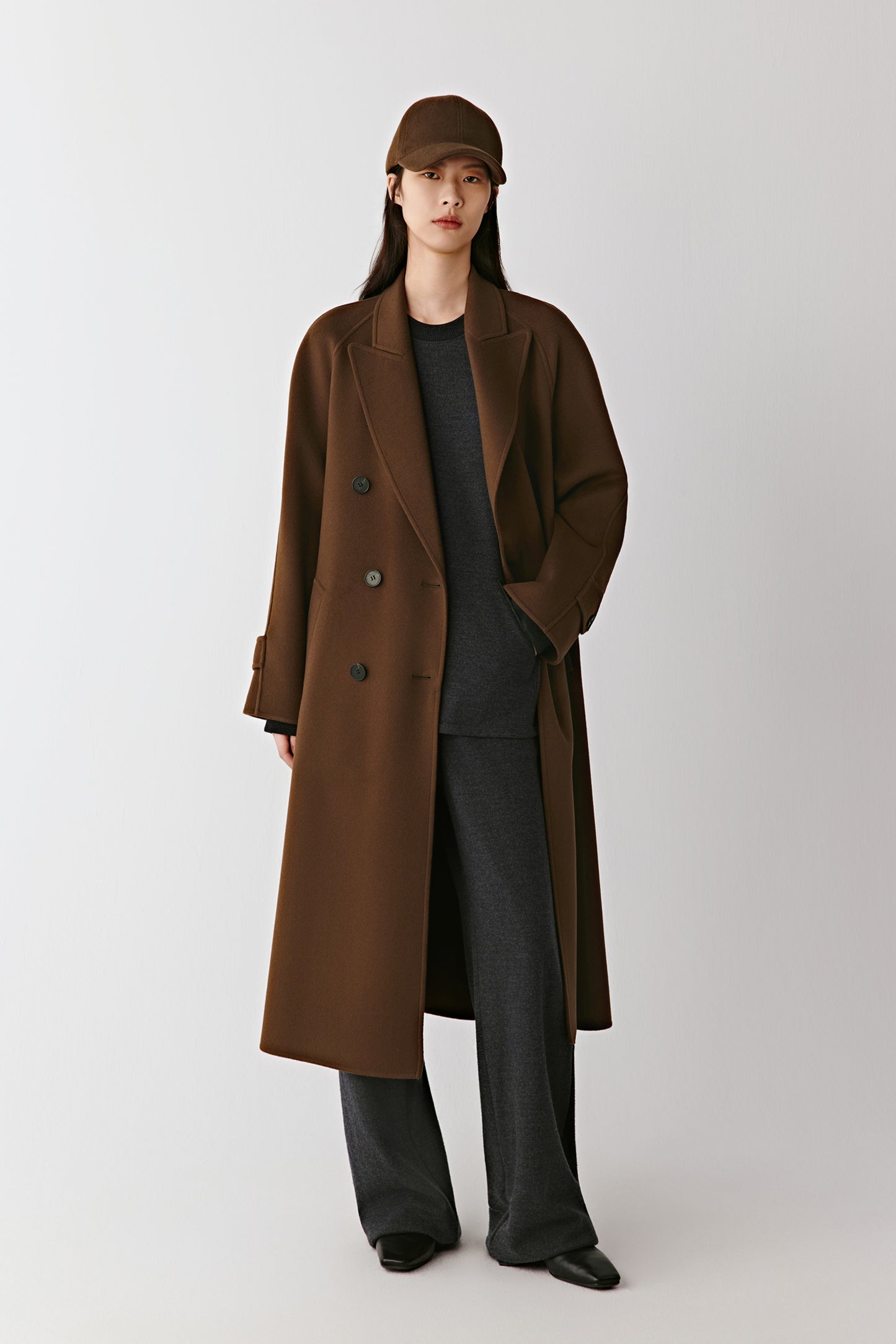 Full-length front view of the model wearing the brown double-faced wool and cashmere coat, showing its long, fluid silhouette and tonal belt.