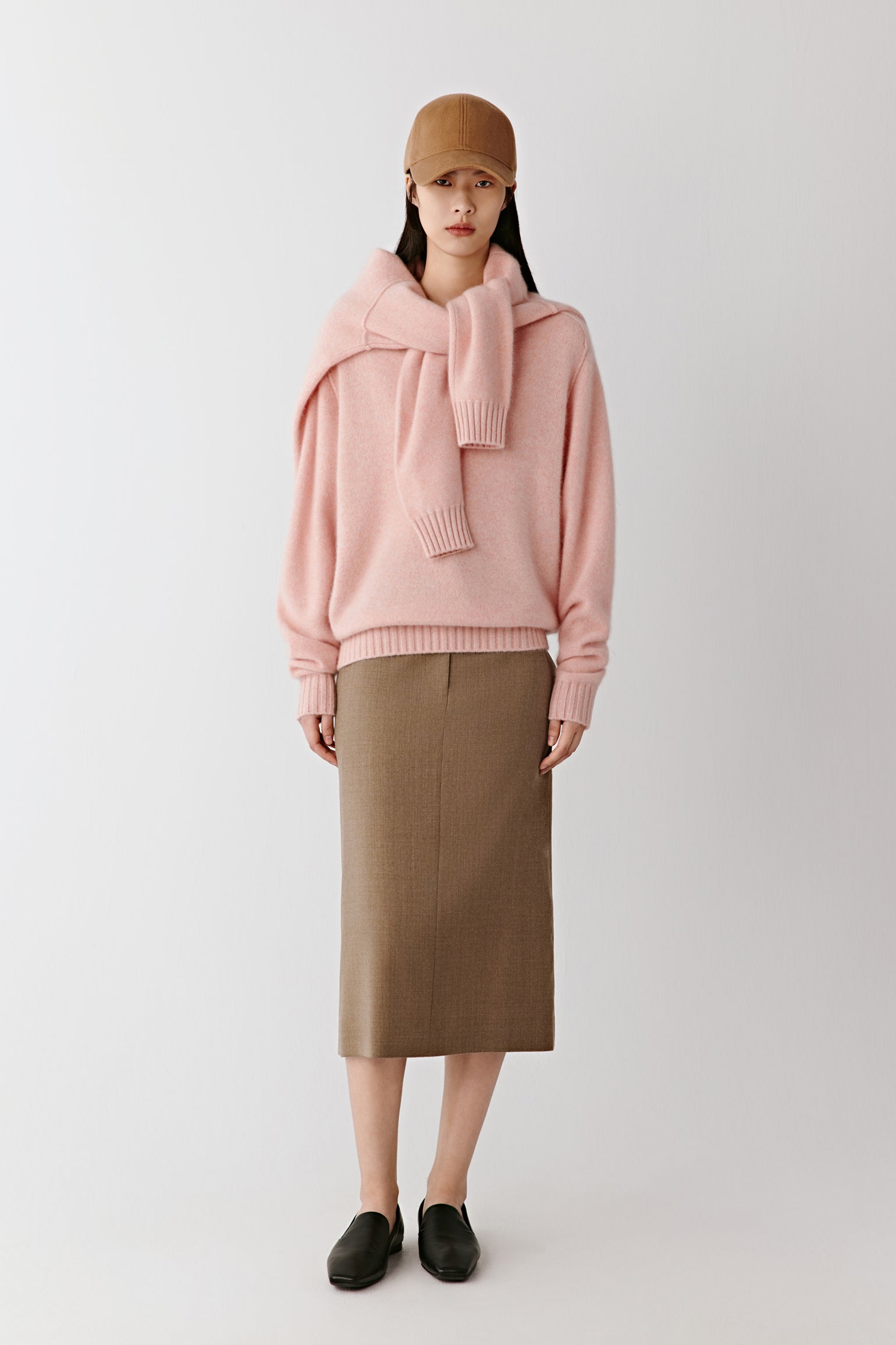 A woman stands facing forward wearing a soft pink cashmere jumper with a V-neck and ribbed cuffs, paired with a brown midi skirt and loafers under soft studio light.