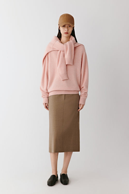 A woman stands facing forward wearing a soft pink cashmere jumper with a V-neck and ribbed cuffs, paired with a brown midi skirt and loafers under soft studio light.
