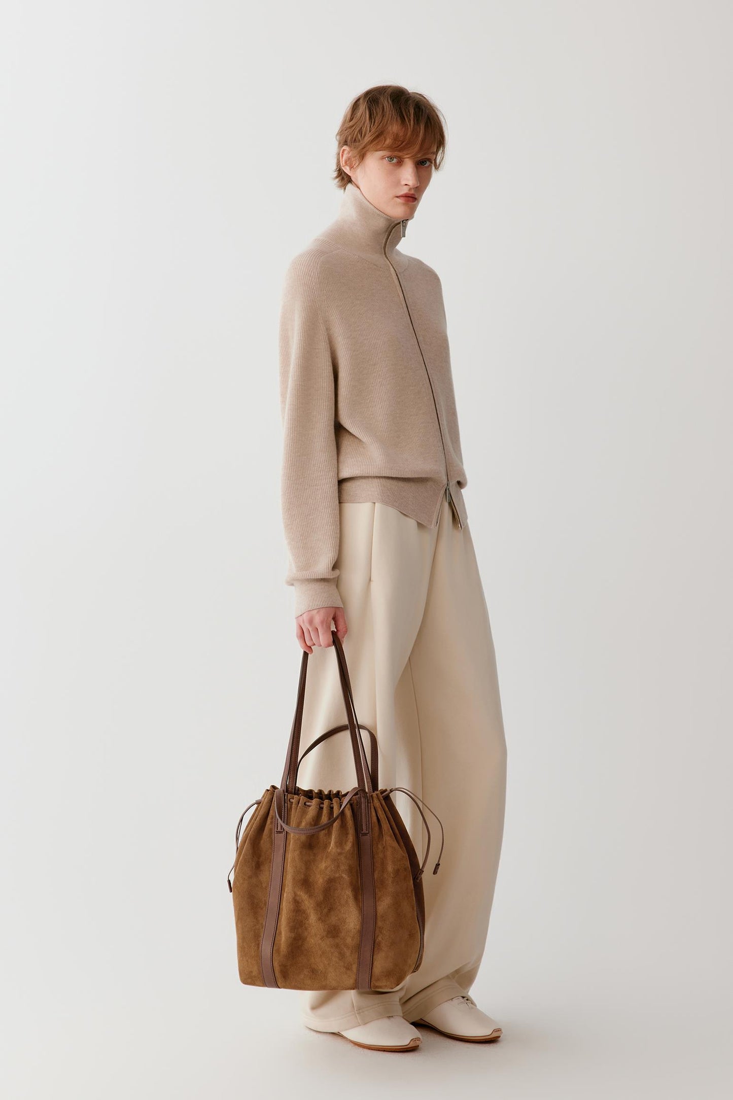 A model stands in profile wearing a beige mock-neck knit top, cream wide-leg trousers, and holding a brown suede drawstring tote bag with dark brown leather handles against a plain light gray studio background.