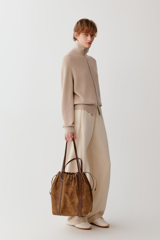 A model stands in profile wearing a beige mock-neck knit top, cream wide-leg trousers, and holding a brown suede drawstring tote bag with dark brown leather handles against a plain light gray studio background.