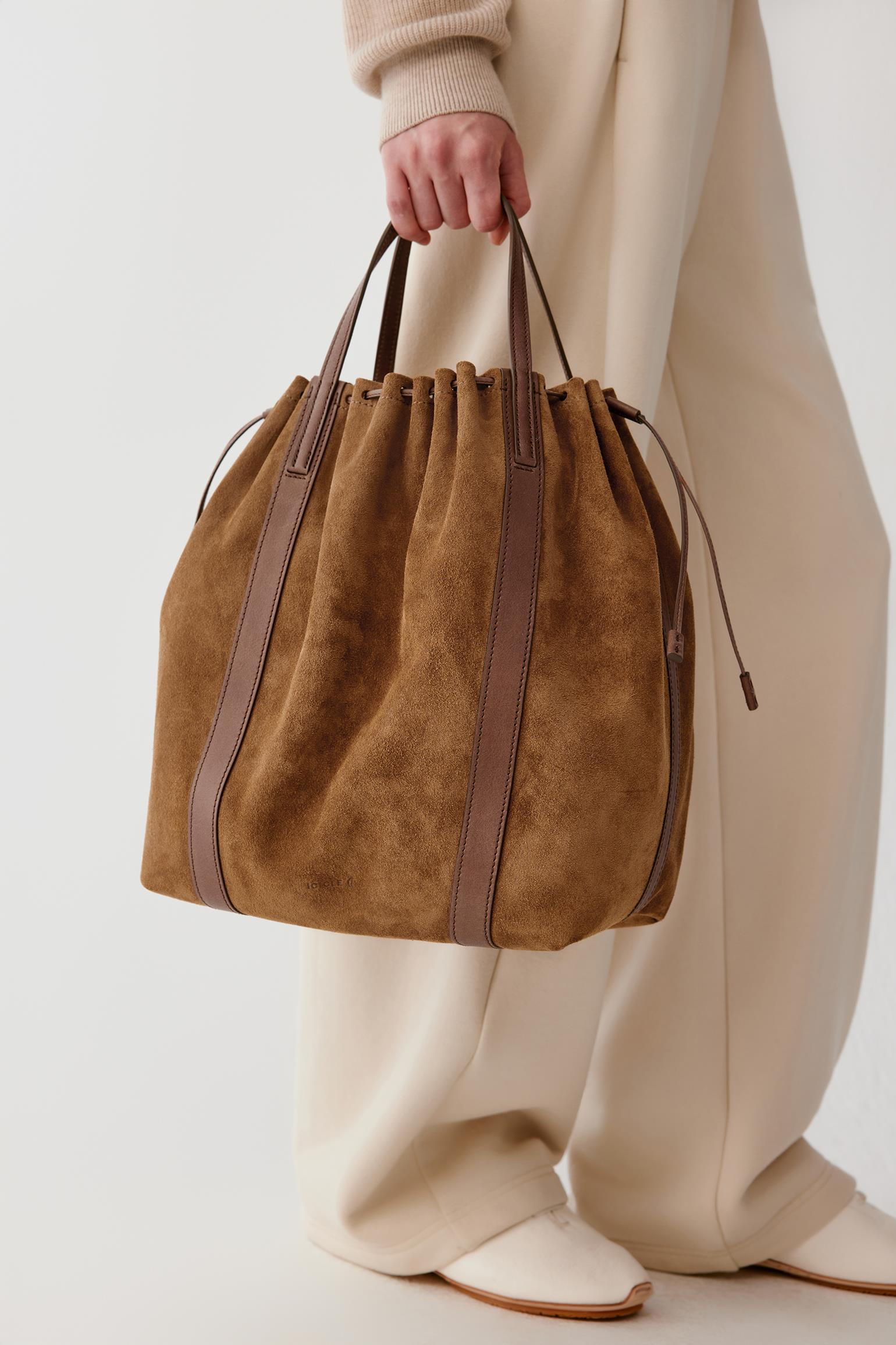 A person in cream-colored clothing holds a brown suede bucket bag with dark brown leather trim and drawstring handles against a plain light background.