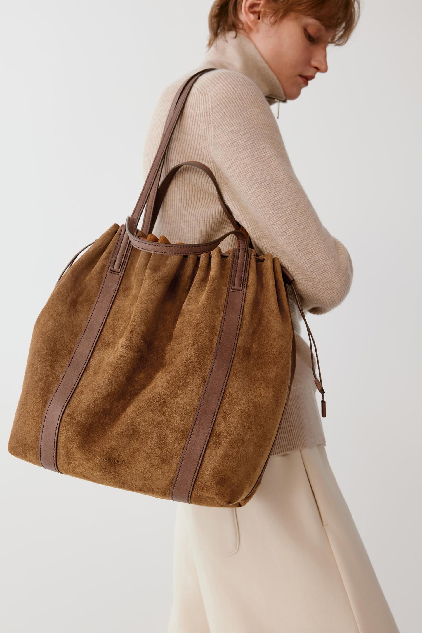A woman wearing a beige ribbed turtleneck sweater and cream skirt carries a large brown suede bucket bag with brown leather straps against a plain light gray background.
