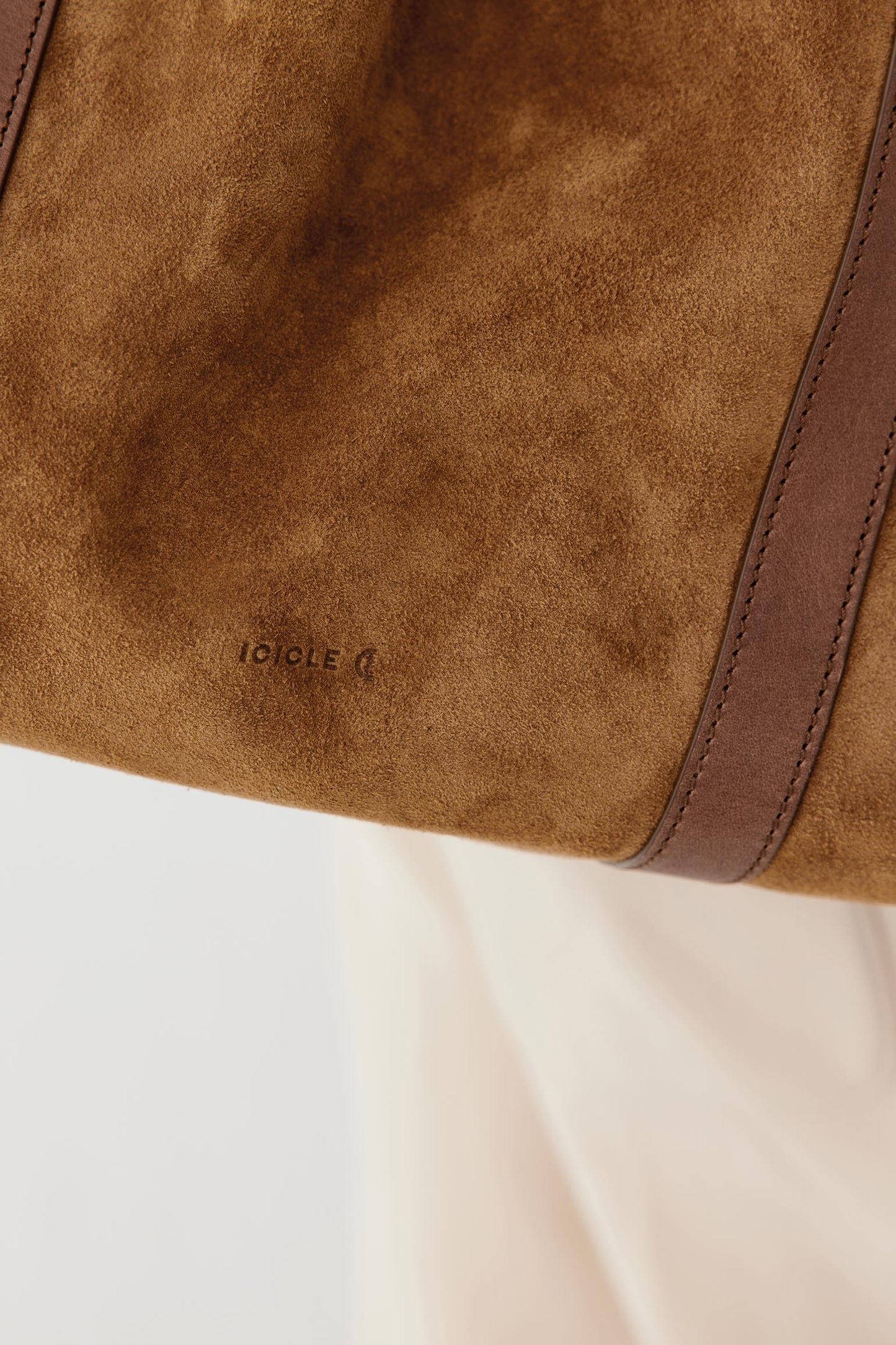 Close-up of a brown suede or suede-like bag surface with a darker brown leather trim strip and a small embossed brand mark reading 'ICICLE'.