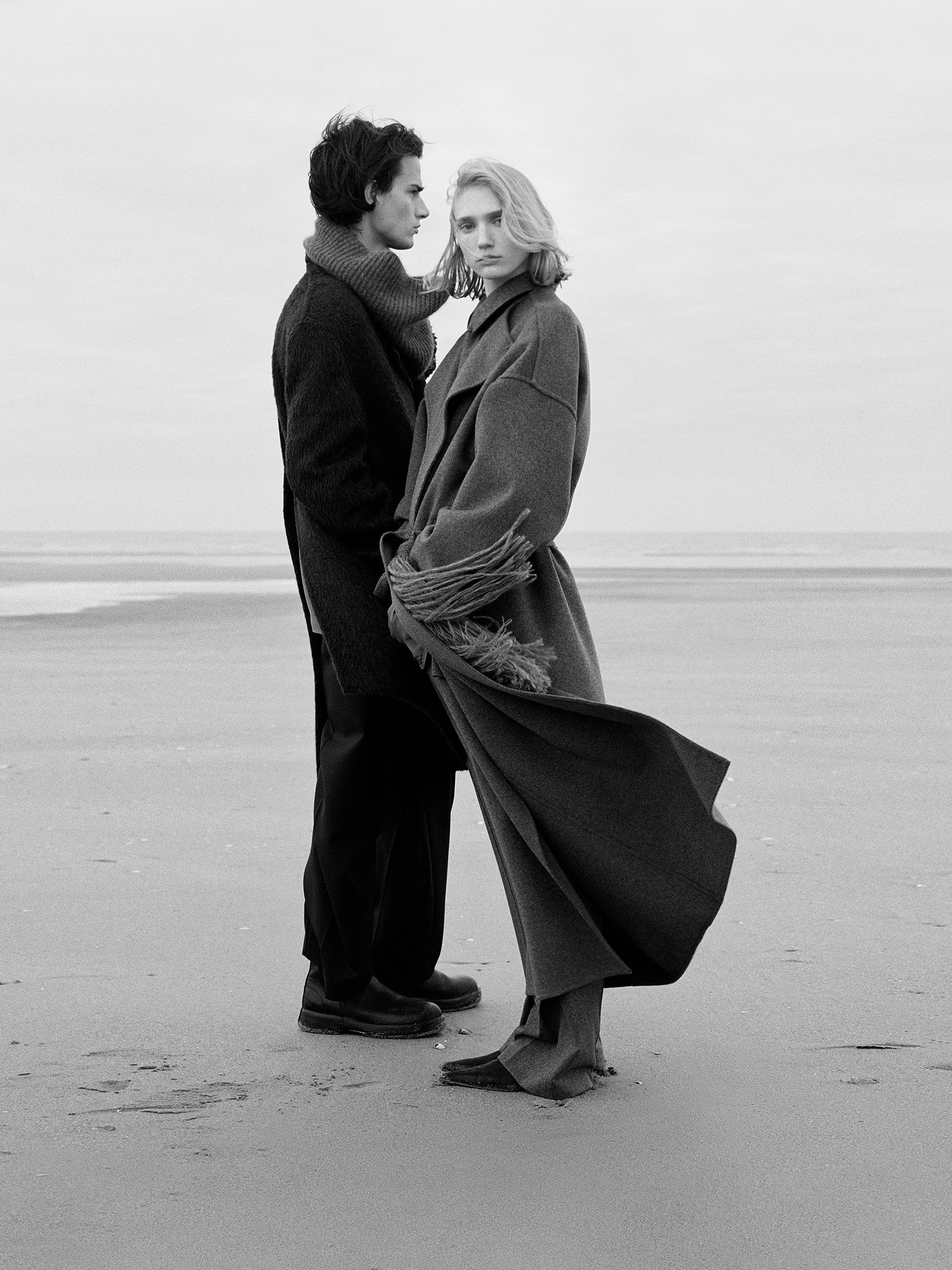 Black and white photograph of two models standing close together on a windswept beach. The man wears a dark turtleneck knit and tailored trousers; the woman faces the camera in a voluminous floor-length wool coat with wide lapels and a fringed scarf wrapped around the arms, coat billowing in the wind. AW26 campaign.