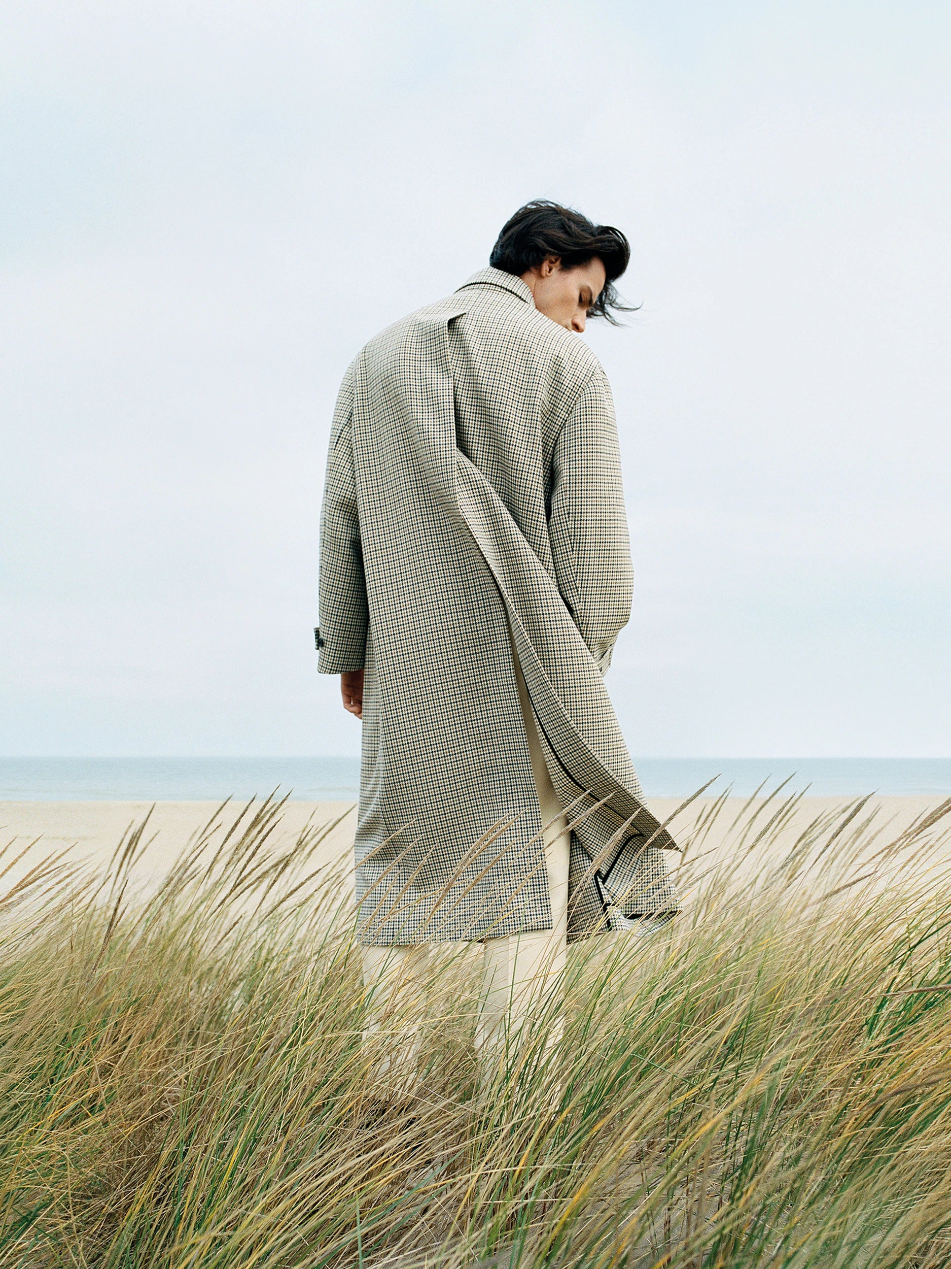 Model seen from behind standing among windswept coastal dunes, wearing an oversized houndstooth check wool coat in beige and black, midi length, with wide lapels and a back vent, paired with cream trousers. AW26 campaign.