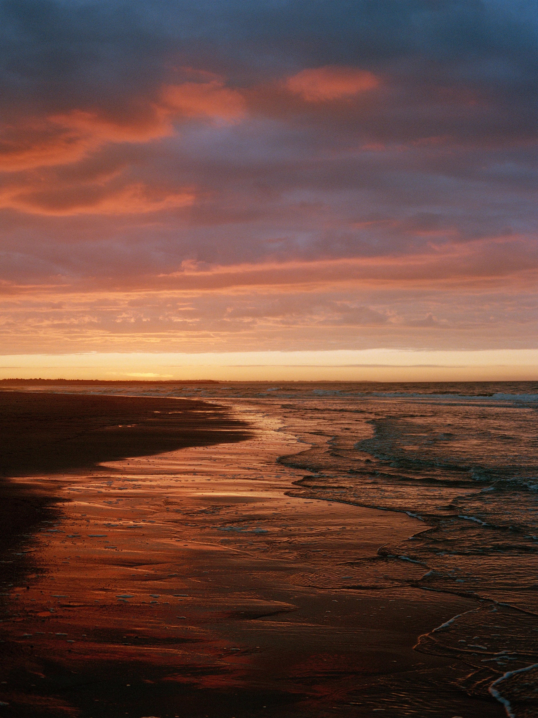 Dramatic sunset over a wide beach at low tide, sky ablaze with deep orange, coral and violet clouds reflected on the wet sand, gentle waves lapping the shoreline. AW26 campaign.