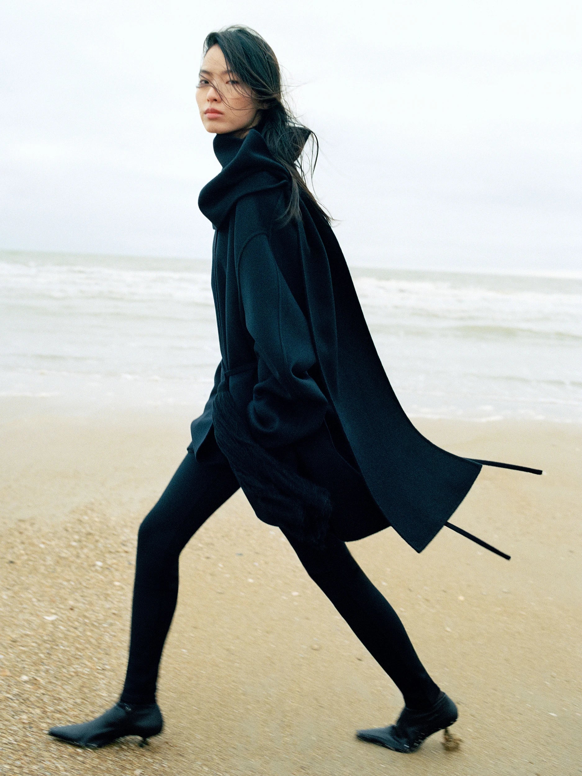 Model walking along a misty beach shoreline, glancing back over her shoulder. She wears an oversized dark navy wool coat with a high draped collar, black leggings and low kitten-heel mules, coat panels trailing in the wind. AW26 campaign.