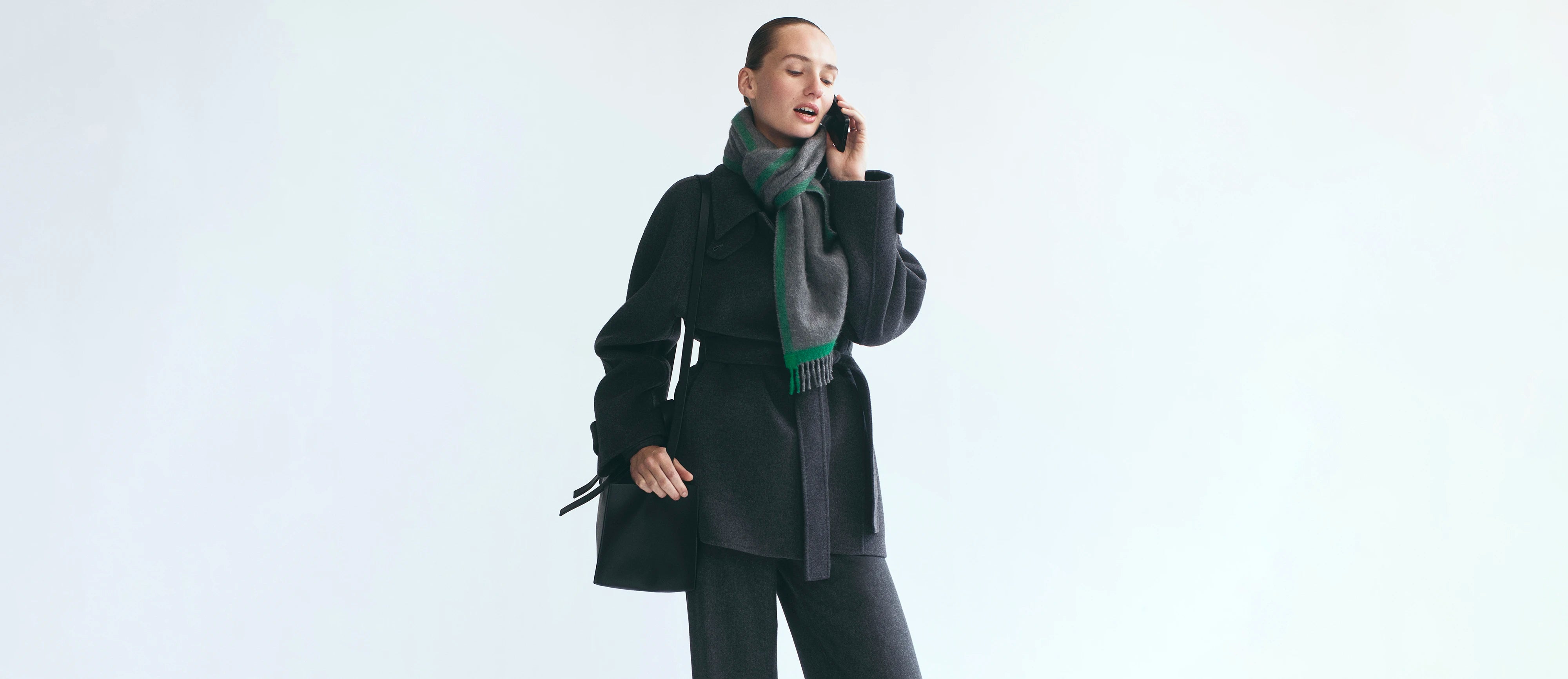 A woman wearing a black coat and a green and gray striped scarf is shown from the waist up, holding a phone to her ear against a plain light gray background.