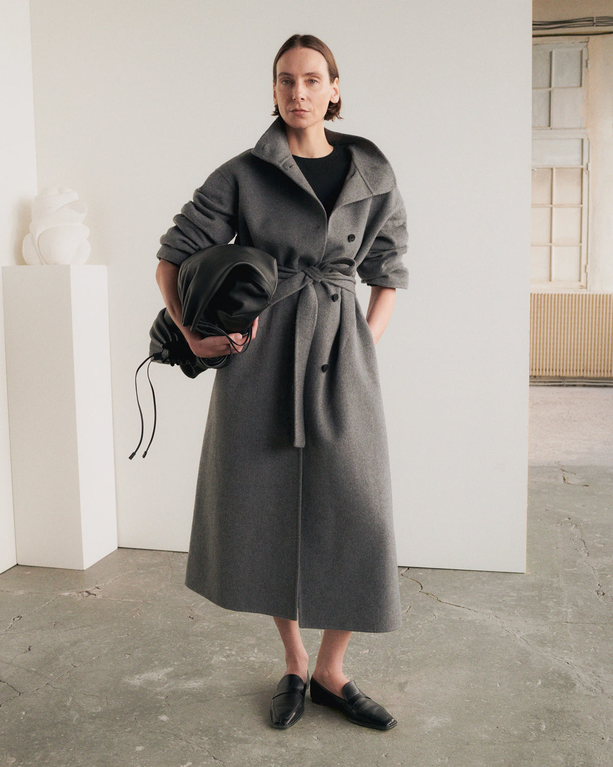 Model standing in a minimalist gallery space with white walls and a concrete floor, wearing an oversized dark grey wool belted coat with wide lapels, three-button front and rolled-up sleeves, tied at the waist with a self-fabric sash, carrying a black leather drawstring pouch, paired with black square-toe loafers. AW26 Icicle A-Line.