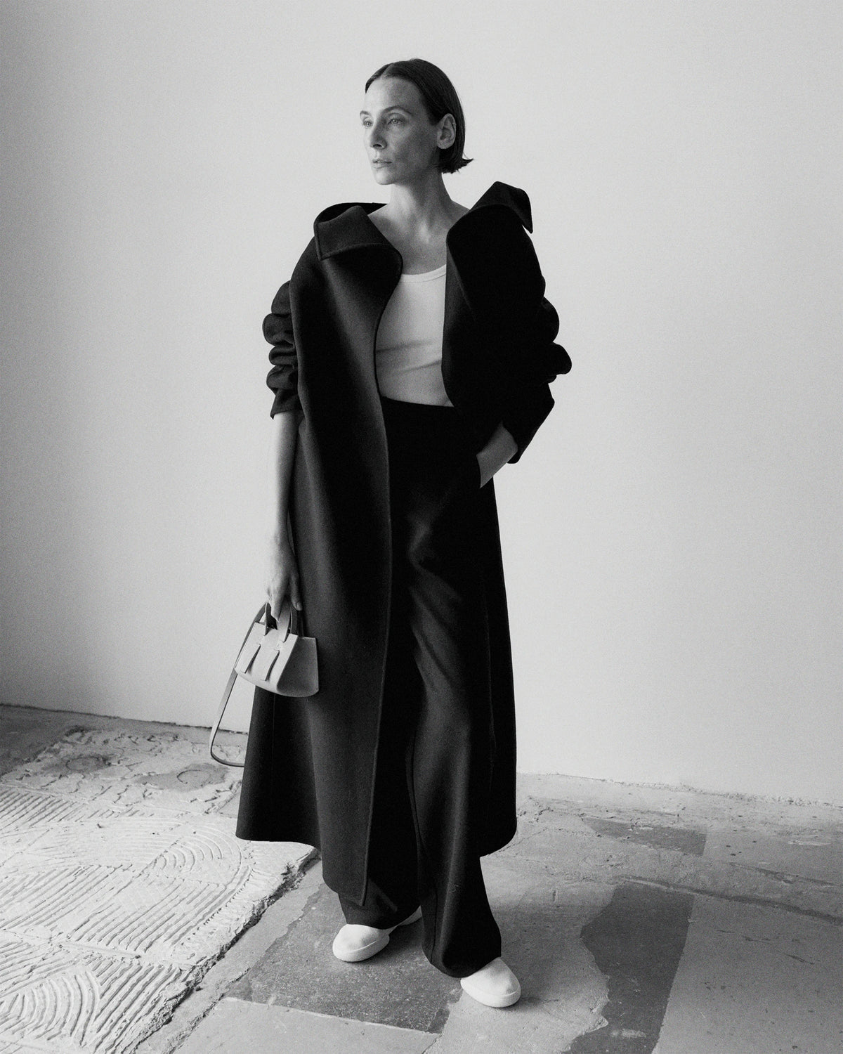 Black and white photograph of a model standing in a minimalist gallery space, wearing a floor-length dark wool coat open over a white fitted top and wide-leg trousers, the coat's shoulders featuring dramatic sculptural ruched detailing, carrying a structured top-handle bag, with white sneakers. AW26 Icicle A-Line.
