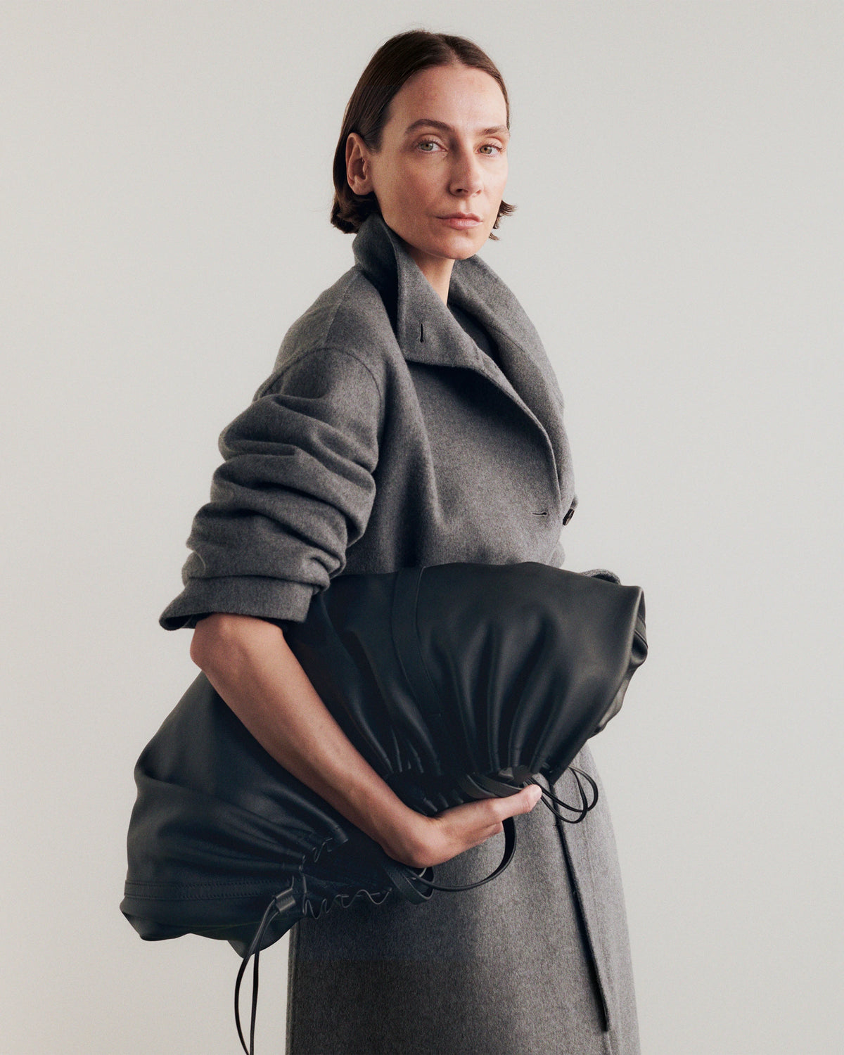 Model in three-quarter profile against a pale grey background, wearing an oversized double-breasted dark grey wool coat with a wide draped collar and voluminous gathered sleeves, holding a large black leather ruched drawstring clutch. AW26 Icicle A-Line.