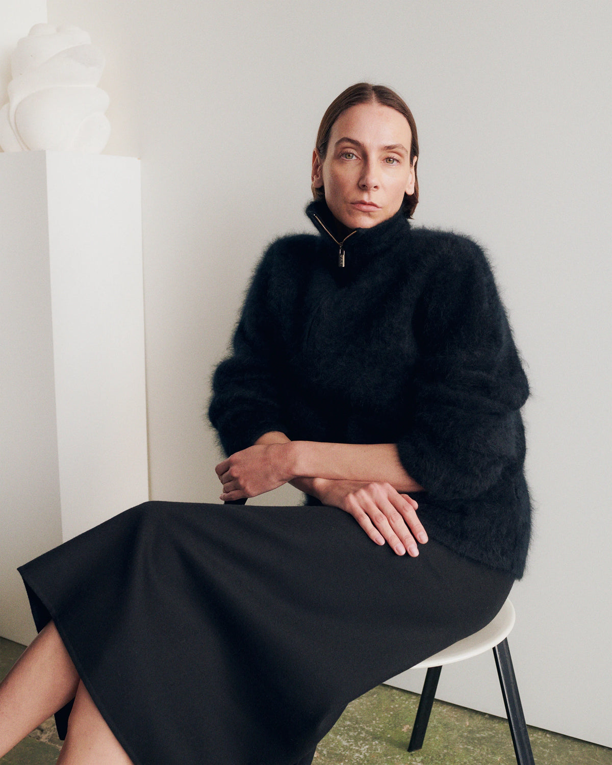 Model seated on a white chair in a minimalist gallery interior, wearing a black fluffy mohair zip-neck sweater with a high funnel collar and ribbed texture, paired with a black wool midi A-line skirt with a side slit. AW26 Icicle A-Line.