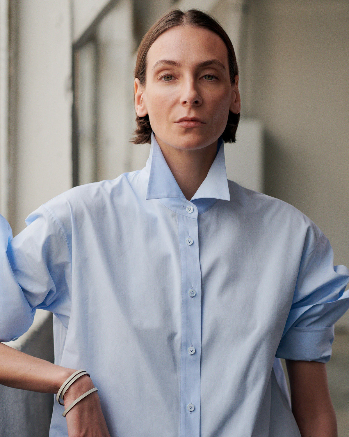Close-up portrait of a model in front of tall windows, wearing an oversized pale blue cotton poplin shirt with a wide spread collar, button front and rolled-up cuffs, accessorised with thin black and white striped bangles on the wrist. AW26 Icicle A-Line.