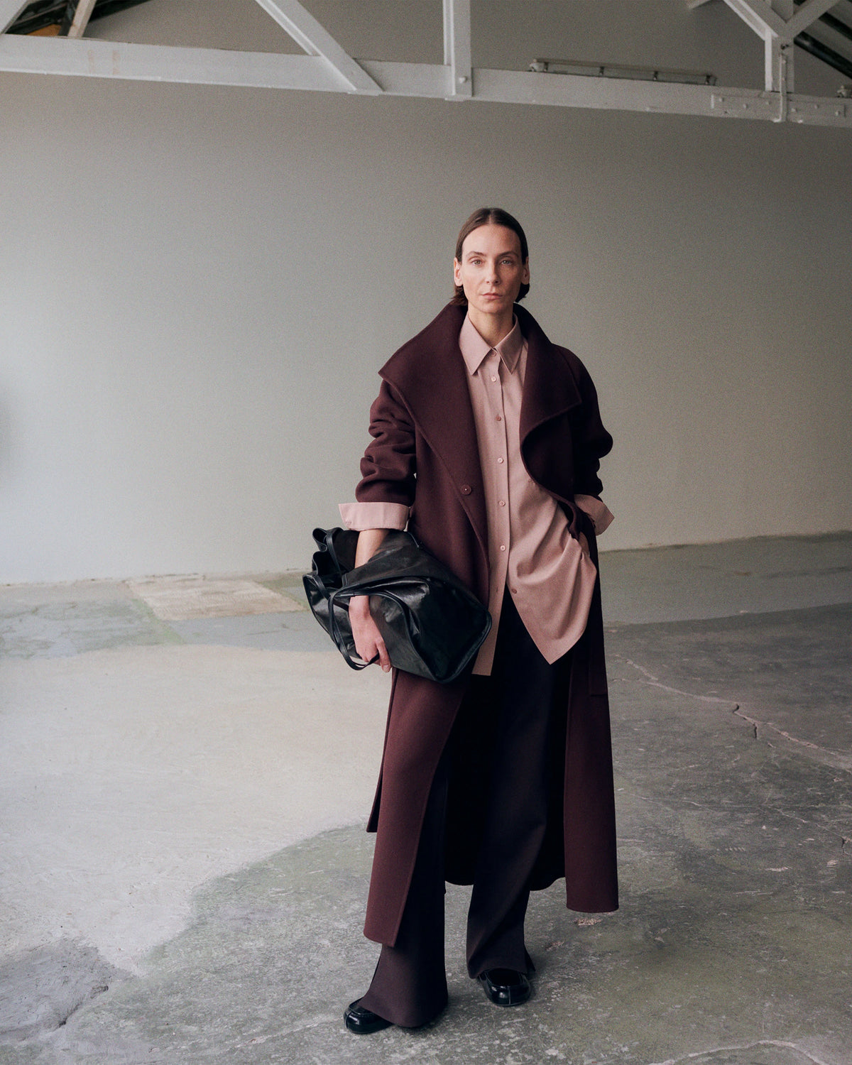 Model standing in a raw concrete industrial space, wearing a floor-length dark burgundy wool coat with an oversized wide lapel collar, open over a loose blush pink button-down shirt, paired with wide-leg black trousers, black patent loafers and sheer black tights, carrying a black leather ruched drawstring clutch. AW26 Icicle A-Line.