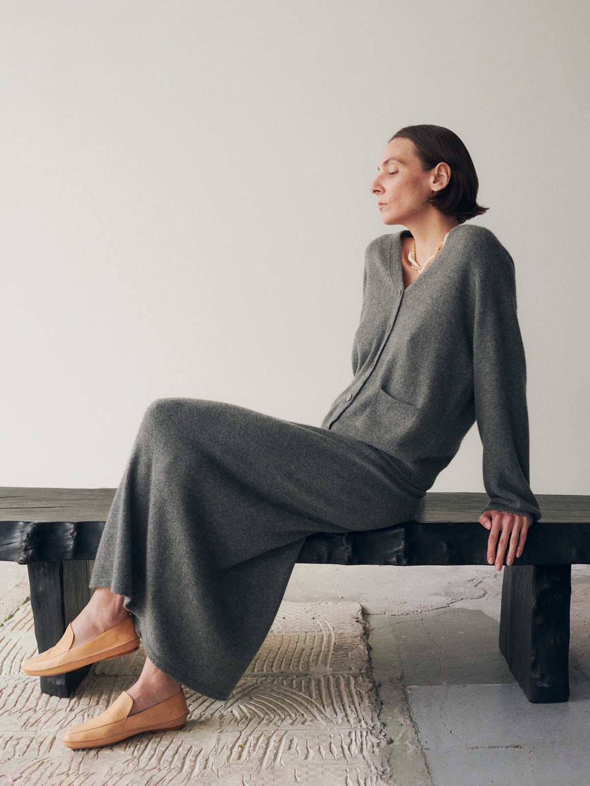 Model seated sideways on a low dark wooden bench with eyes closed, wearing a dark grey marl cashmere V-neck cardigan with button front and a matching floor-length knit skirt, accessorised with a gold sculptural pendant necklace and tan leather penny loafers, on a textured cream woven rug. AW26 Icicle A-Line.