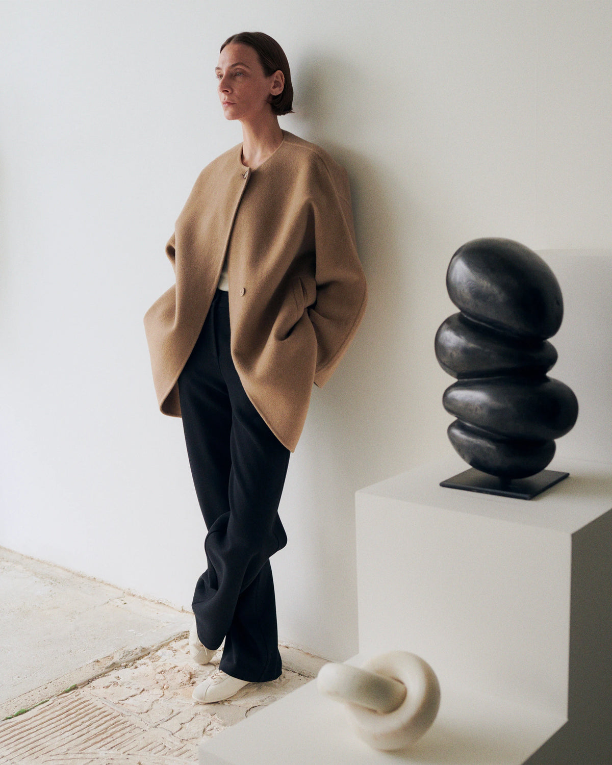 Model leaning against a white wall in a gallery space, gazing to the side, wearing an oversized camel wool collarless jacket with a single button and deep side pockets, paired with wide-leg black trousers and white sneakers; a black abstract stacked stone sculpture on a white plinth stands beside her. AW26 Icicle A-Line.